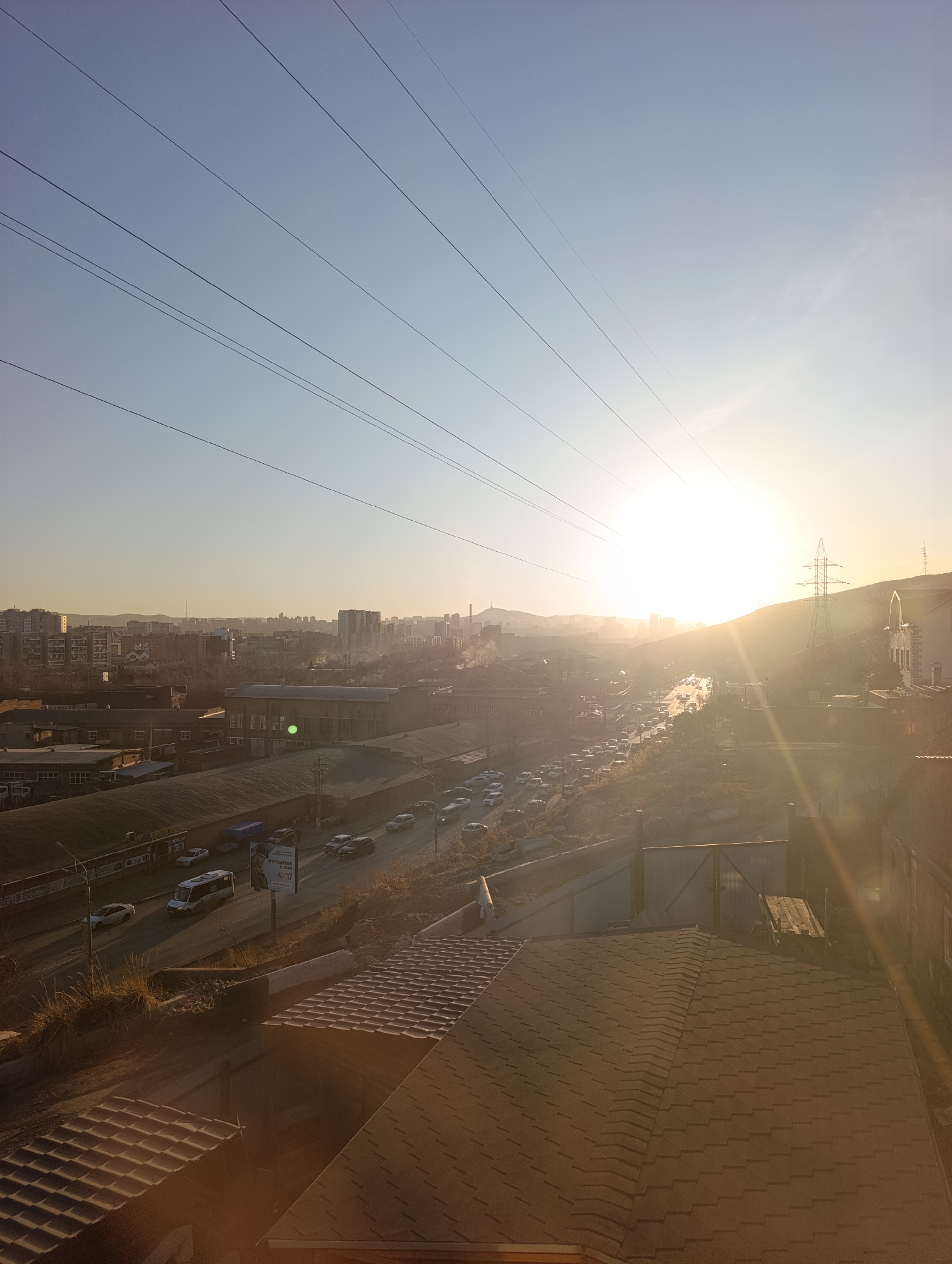 The image shows an elevated view of a cityscape at sunrise or sunset, likely in Krasnoyarsk, Russia. The bright sun obscures details in the center, but power lines stretch across the top of the frame. The city below consists of a mix of industrial buildings and residential structures. A busy road filled with cars is visible, and a hill is situated in the background with a transmission tower. In the foreground, a roof with brown shingles is seen. There is no evidence of people in the image.