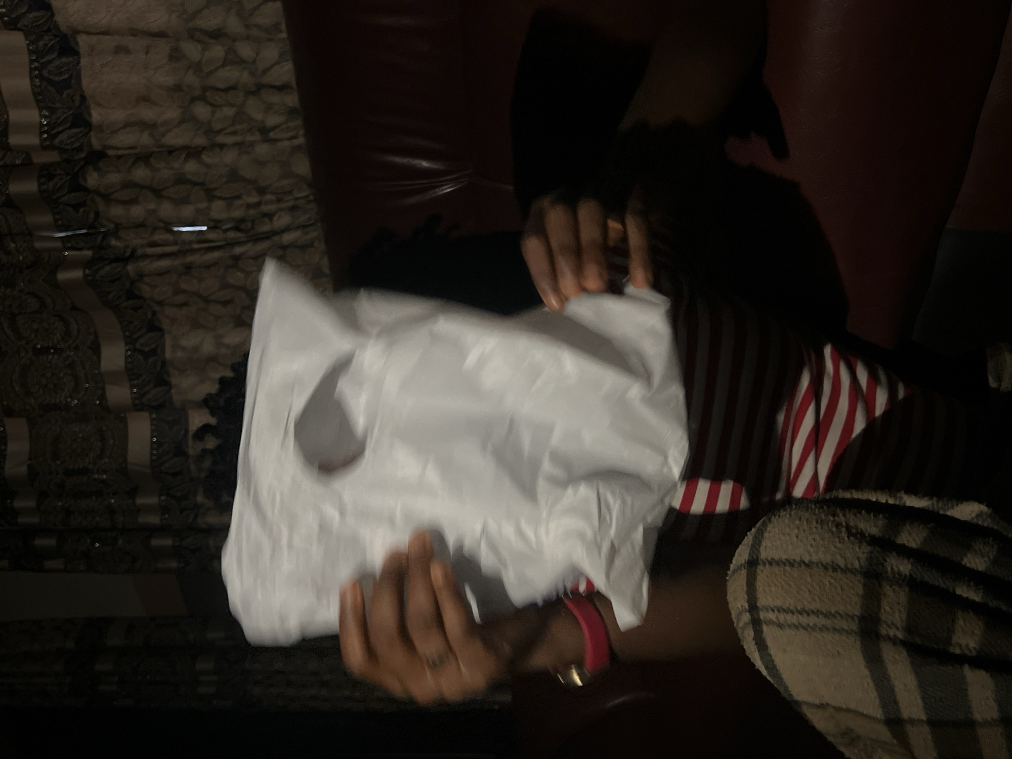 A person is seated on a dark red couch in Nkpor, Nigeria. The individual is holding a white plastic bag in front of their face, obscuring their features. They are wearing a red and white striped shirt and plaid pants. Behind them, dark patterned curtains are visible. The lighting is dim.
