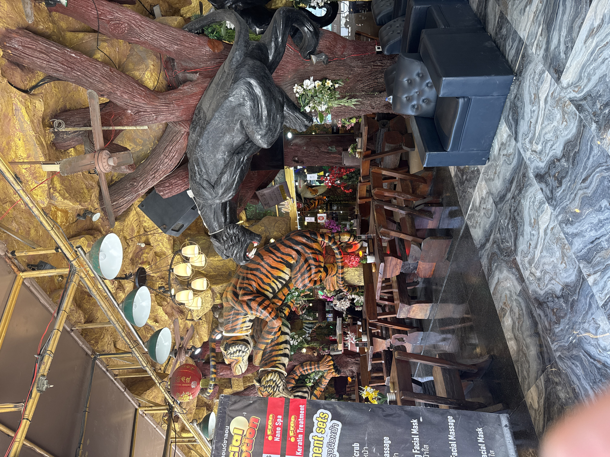 The image depicts the interior of a spa and massage parlor, likely located in Patong, Thailand, given the Thai text visible on a banner. The decor is elaborate and features several large, sculpted tiger figures intertwined with a faux tree trunk that dominates the center of the space. A black panther sculpture is also visible perched on the tree. The seating area includes dark wooden tables and benches, arranged in a rustic style. In the foreground, to the right, are modern black leather armchairs. The ceiling is adorned with decorative lighting fixtures, including a red lantern and several pendant lights. The walls are textured to resemble rock formations and are painted in gold and yellow hues. The floor is a polished marble with a swirled pattern. The overall impression is one of a uniquely themed, somewhat kitschy, establishment designed for relaxation and spa services. There are no people visible in the scene.