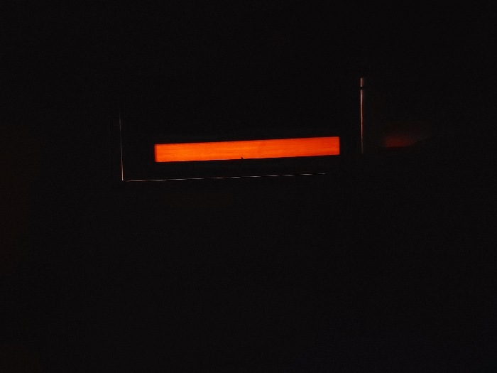 The scene, located in Chaikovsky, Russia, depicts a predominantly dark interior environment. A vertical, rectangular strip of intense orange light is positioned on the right side of the frame, occupying approximately one-fifth of its width. This light is contained within a subtle, dark rectangular boundary, indicating it originates from a narrow aperture or an integrated light fixture. Below the primary orange light, a fainter, diffused orange-brown glow is discernible on a lower, undifferentiated surface, consistent with light spill. No individuals, additional objects, or explicit actions are visible within the dark surroundings.