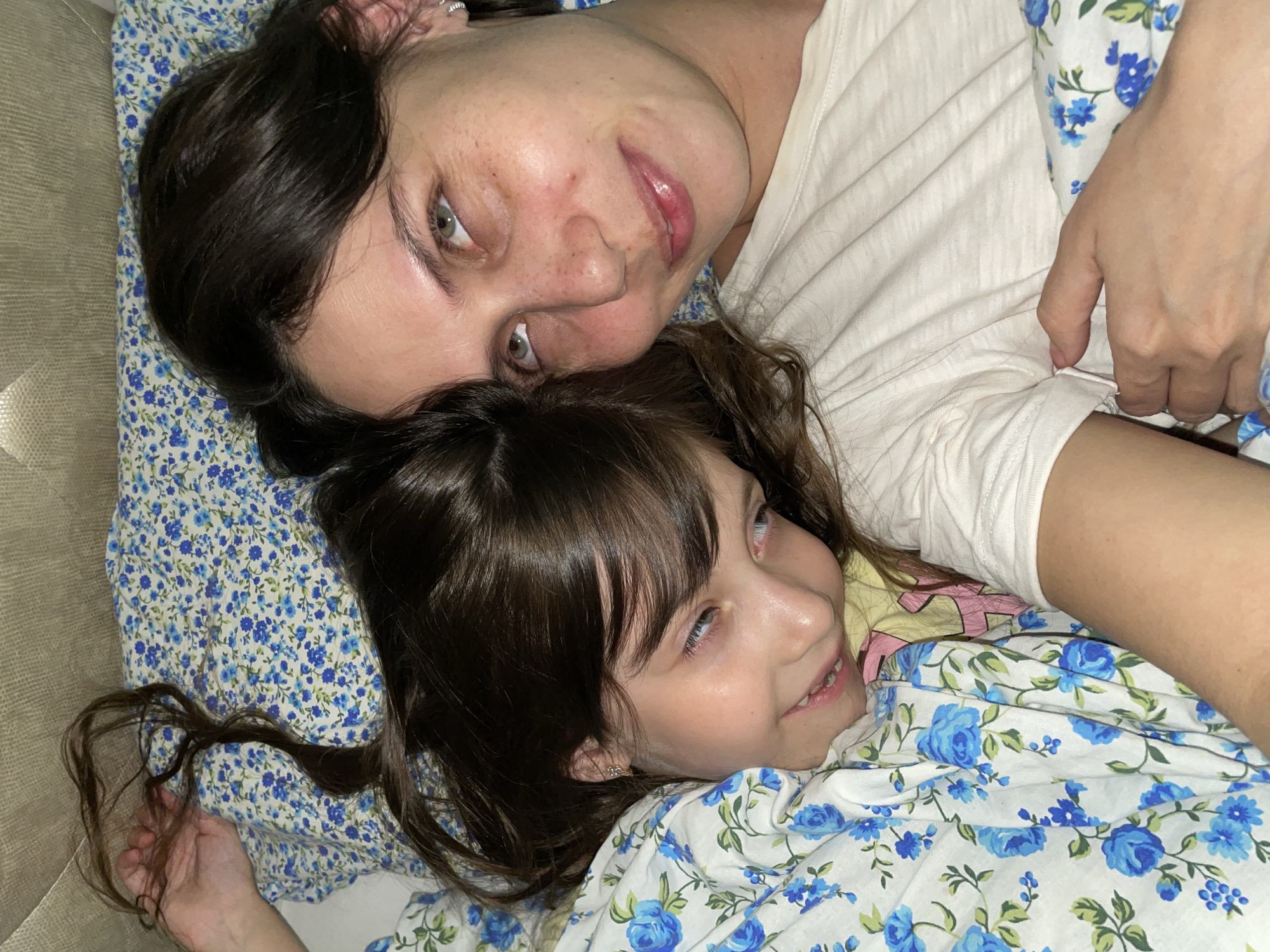 This media file depicts an intimate overhead view of two females, an adult and a child, likely a mother and daughter, lying closely together indoors. Both individuals have dark brown hair. The adult is positioned above and behind the child, with her head resting on a pillow. She has light green or hazel eyes and a subtle, content smile, wearing a plain white t-shirt. The child, nestled below, has blue eyes and a calm, slightly upward gaze, partially covered by a patterned blanket.
The bedding, including pillows and blankets, features a repeating design of blue roses and green leaves on a white background. A neutral-toned surface, possibly a sofa or headboard, is visible along the left edge of the frame. The scene conveys a sense of warmth, relaxation, and affection, suggesting a quiet moment of bonding.
The image is taken from a top-down perspective. There are no clear indications of the time of day or external weather conditions. While the context of Izhevsk, Russia, is provided, there are no specific visual cues within the image to confirm this location. No text is visible in the photograph. The overall mood is peaceful and tender.