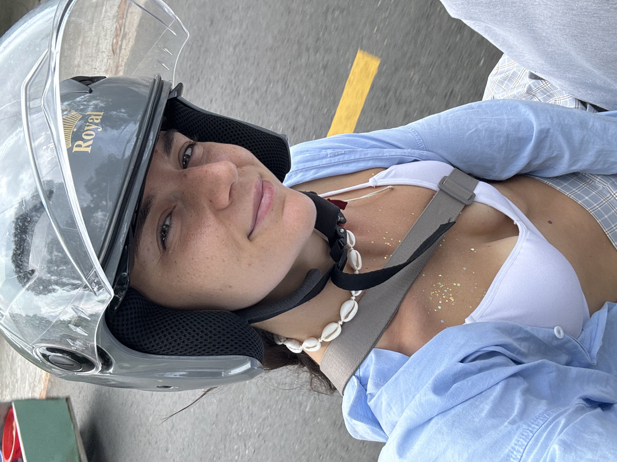 A young Caucasian woman with light skin, freckles, and dark brown hair is shown from the chest up, riding a motorcycle in Mũi Né, Vietnam. She is wearing a grey helmet with a clear visor, a white bikini top, and a light blue open shirt. A seashell necklace is visible around her neck, and gold glitter is applied to her décolletage. She is looking directly at the camera and smiling. The background is blurred, indicating movement, and shows a grey asphalt road with a yellow line. The scene suggests a casual outdoor activity on a bright day.