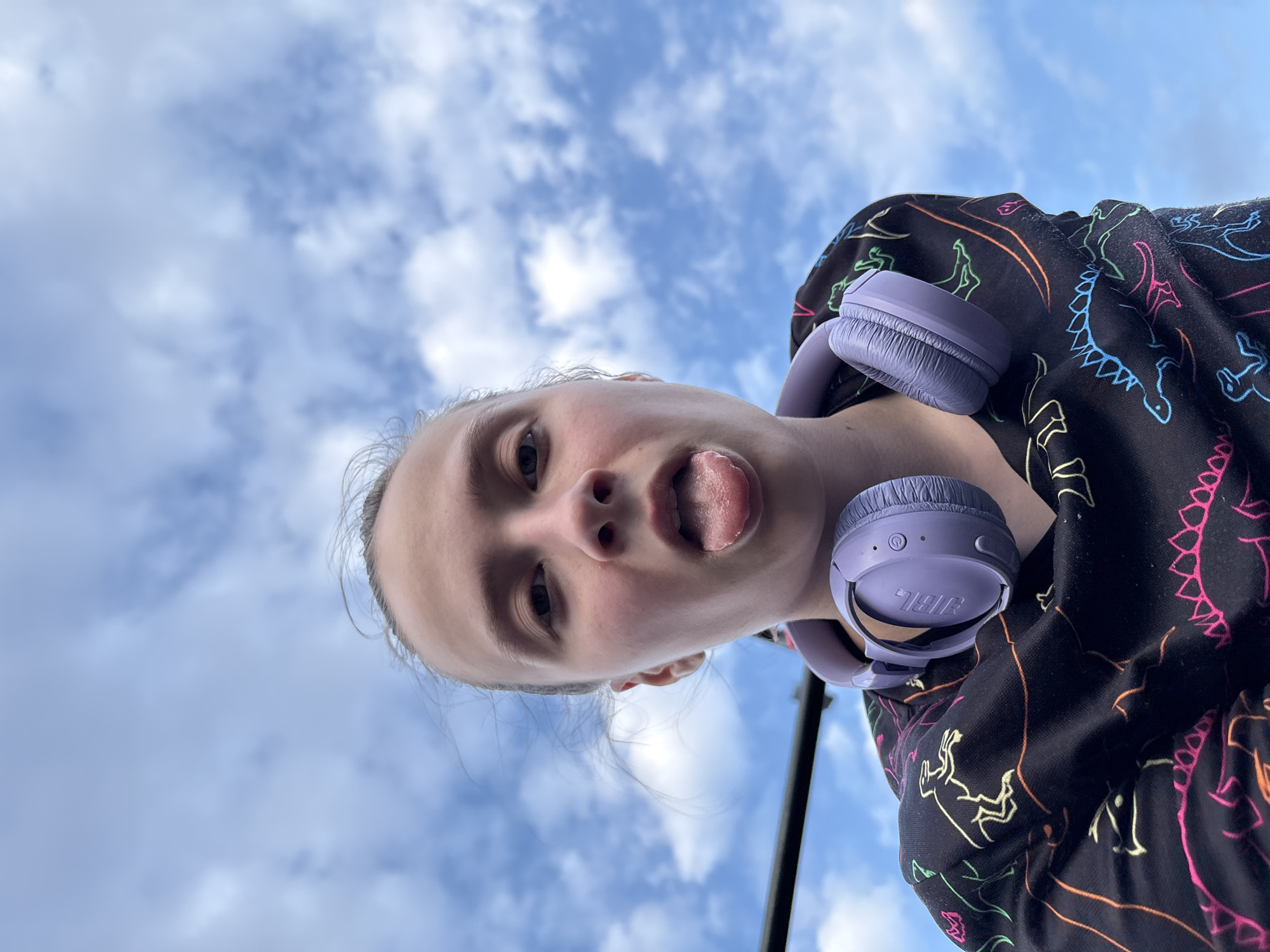 A young person is depicted in a low-angle shot against a sky filled with clouds. They are sticking out their tongue, and purple headphones are positioned around their neck. The person is wearing a dark-colored hoodie with a dinosaur design on it. The picture appears to have been taken outdoors. There are no indicators to discern the city where the photo was taken.
