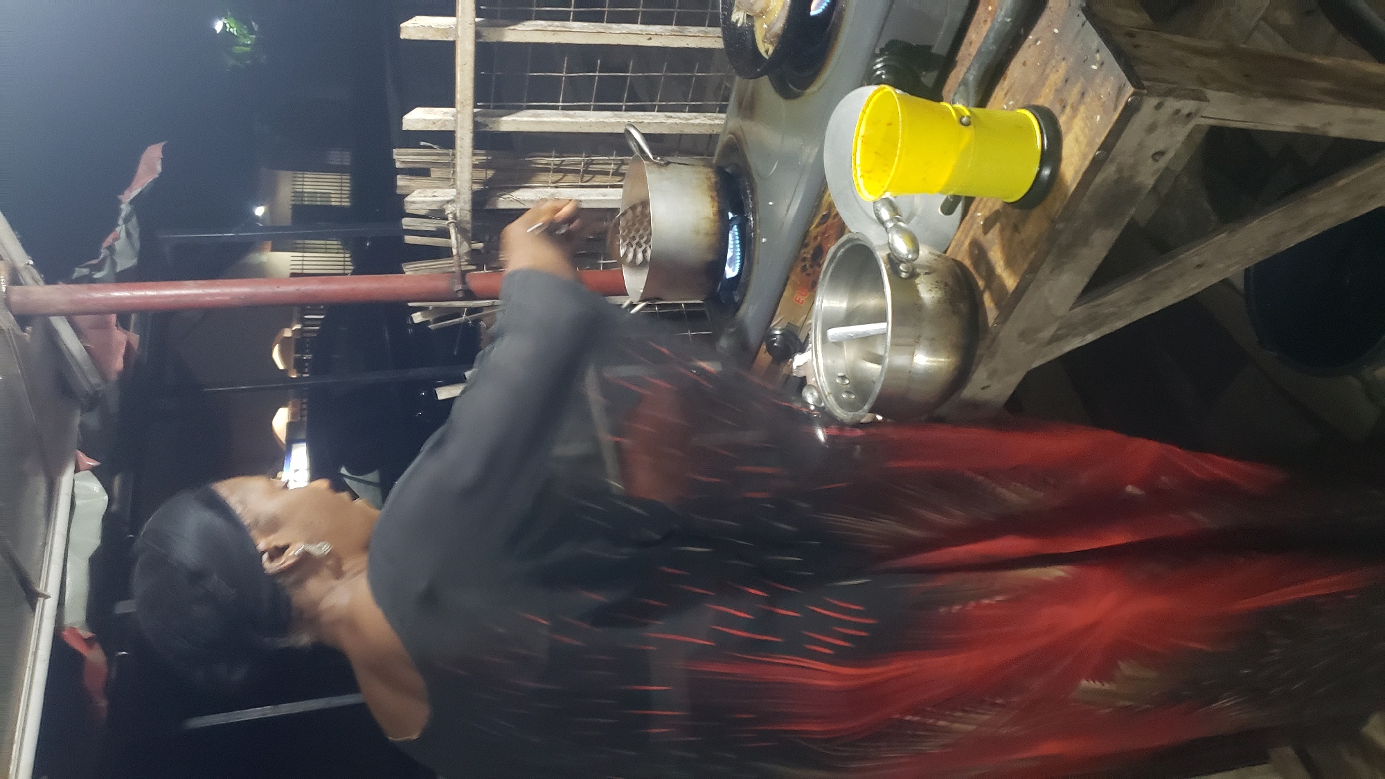 A medium shot shows a Black woman from the chest up, wearing a black headscarf and a black and red patterned dress, standing at a food stall in Narayi, Nigeria. The woman is facing right, with her head tilted down towards a stove. She is stirring a pot on the stove with a slotted spoon. The stove is a gray, two-burner gas stove. A second pot sits on a lower shelf next to the stove. To the right of the stove is a yellow plastic bucket. The stall is covered by a makeshift roof made of corrugated metal and plastic sheeting. A wooden table is in the foreground. The background is dark, with some indistinct lights visible in the distance. The scene appears to be at night.