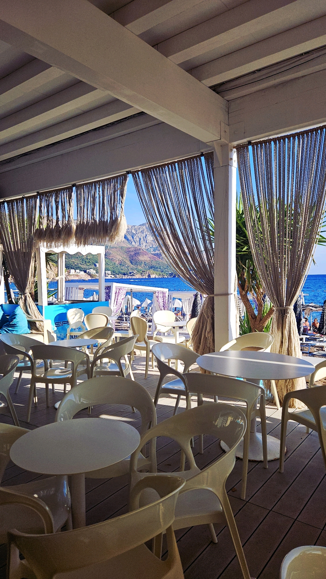 The image depicts an outdoor cafe with a view of the ocean and mountains. White tables and chairs are arranged on a wooden deck, with rope curtains hanging from a white overhead structure. In the background, the blue ocean stretches out to the horizon, meeting a mountainous coastline under a clear sky. Several umbrellas and lounge chairs are visible on a beach in the distance, and a few people are present. The overall atmosphere is bright and sunny, suggesting a pleasant day at a seaside resort.