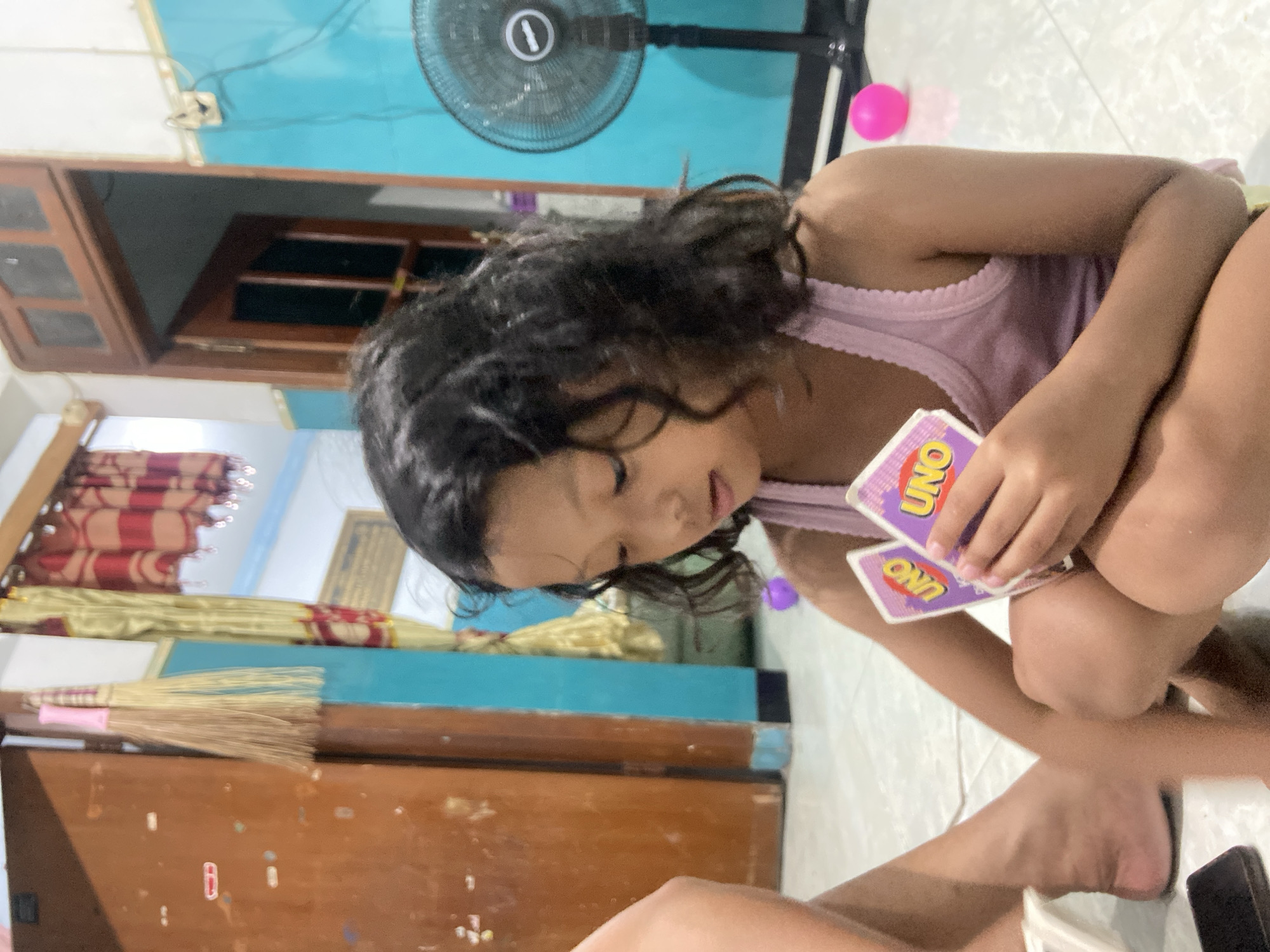 A young Indonesian girl is sitting on a tiled floor in what appears to be a home in Gelam, Indonesia. She is holding a few Uno cards in her hands and is looking down at them. She has long, dark, curly hair and is wearing a light pink tank top. Her left leg is bent, and her right leg is tucked underneath her. To her left, another person's legs are visible, indicating they are also present in the scene. The background shows a wooden door, a blue wall with some electrical outlets, and a black electric fan. There are also some curtains hanging from the ceiling and a broom leaning against the wall. A pink ball is visible on the floor behind the girl.