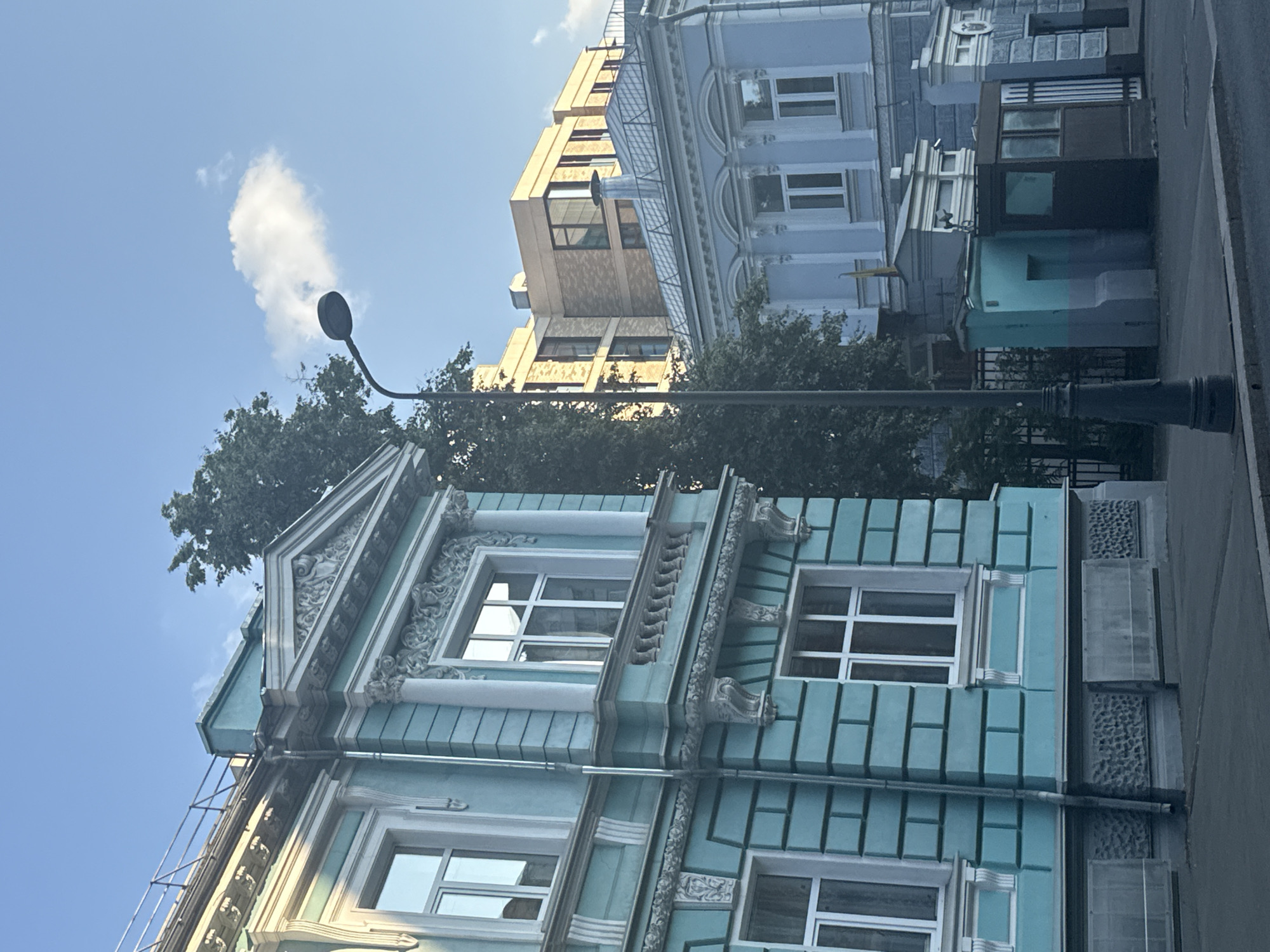The image depicts a street scene in Moscow, Russia, focusing on the architecture of several buildings. In the foreground, a light blue building with ornate white detailing and numerous windows stands prominently. The building features decorative friezes, cornices, and pilasters, suggesting historical architecture. Adjacent to it, a taller, modern yellow building with large windows rises. Further back, a pale blue building with arched window frames and classical architectural elements is visible. The scene is illuminated by natural light, indicating it is daytime. The sky is clear with a few white clouds, suggesting fair weather. A streetlamp with a curved arm is visible in the mid-ground, and lush green trees partially obscure some of the buildings. The perspective is angled upwards, emphasizing the height and details of the structures. The image conveys a sense of urban architecture, likely in a historic part of Moscow. No people or specific activities are discernible. There is no visible text in the image.