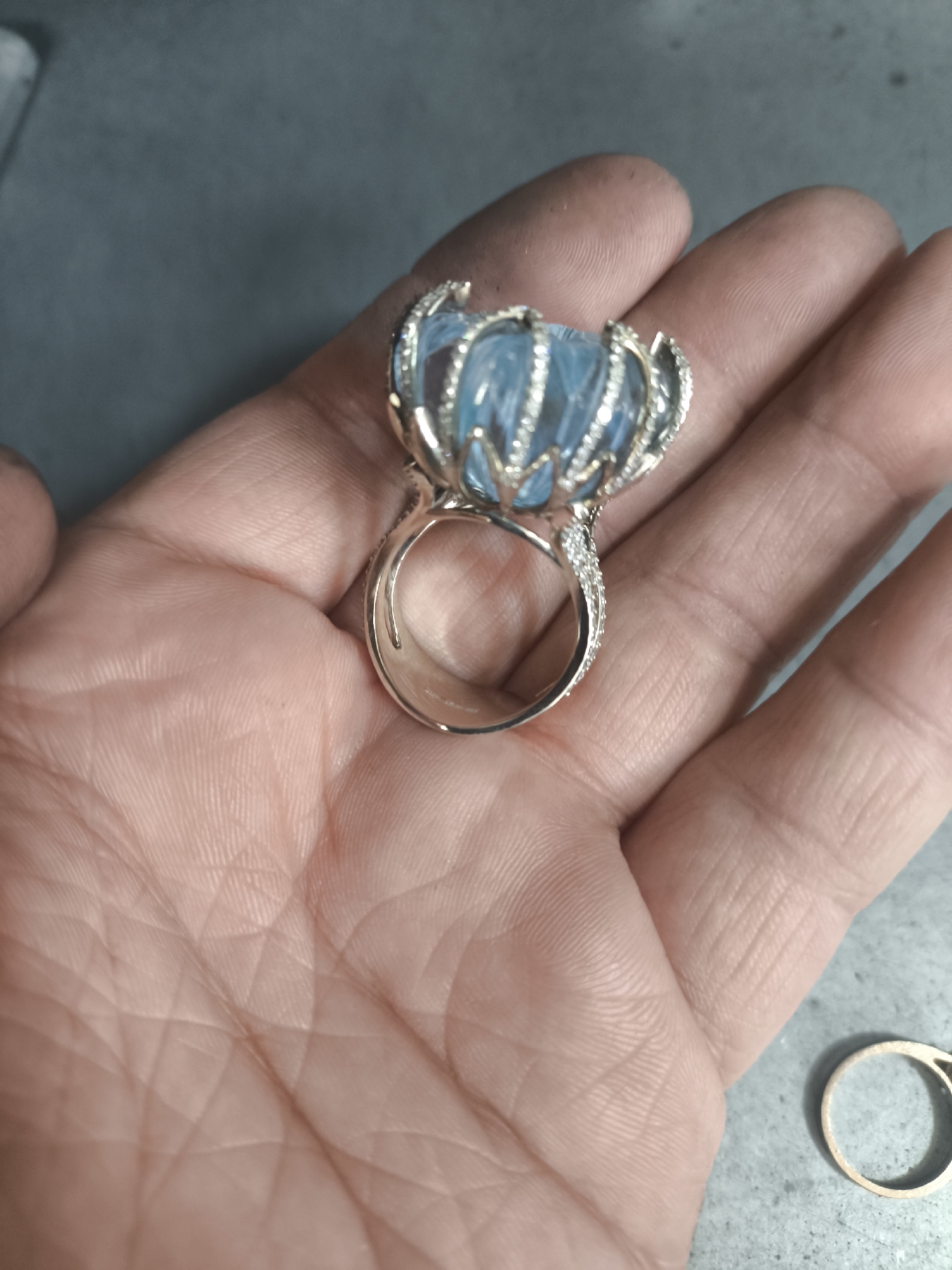 A close-up, top-down, angled shot shows a person's open palm, with a large, ornate ring resting on it. The ring features a prominent, faceted, light blue gemstone, possibly aquamarine, that appears to be carved in a floral or crown-like design. The setting of the ring is silver or white gold, encrusted with small, clear stones, likely diamonds, along the band and the prongs holding the main gemstone. The hand shows visible lines and texture, with a slight dusting of what appears to be fine particulate matter on the skin and the ring setting. In the lower right corner of the frame, another plain gold ring is visible on a grey, textured surface. The background is a muted, greyish surface. There is no indication of the city of Sredneuralsk, Russia, or any other specific geographical location.