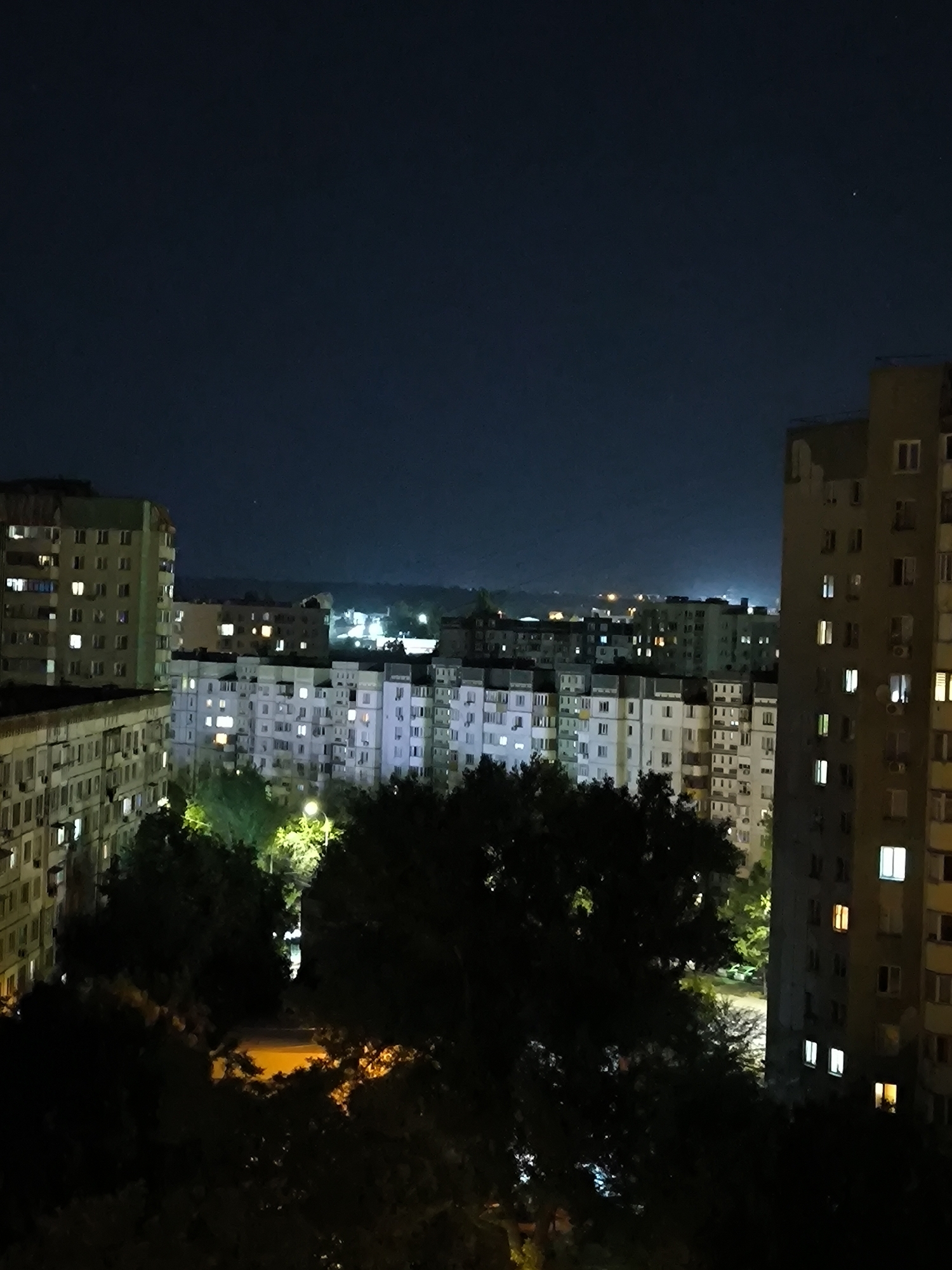 This is a nighttime, wide-angle, eye-level, outdoor, full shot of a residential area in Chișinău, Moldova. The sky is dark blue with a few visible stars and a faint haze on the horizon. In the foreground, silhouetted trees obscure the lower part of the frame. Behind the trees, several multi-story apartment buildings are visible, with many windows illuminated, indicating occupancy. The buildings are of varying heights and architectural styles, typical of Soviet-era construction. One prominent building on the far right is a tall, grey structure with uniformly lit windows. To the left, another building is a lighter color with green trim. In the background, more buildings and distant lights suggest a sprawling urban landscape. The overall scene depicts a quiet, illuminated city at night.