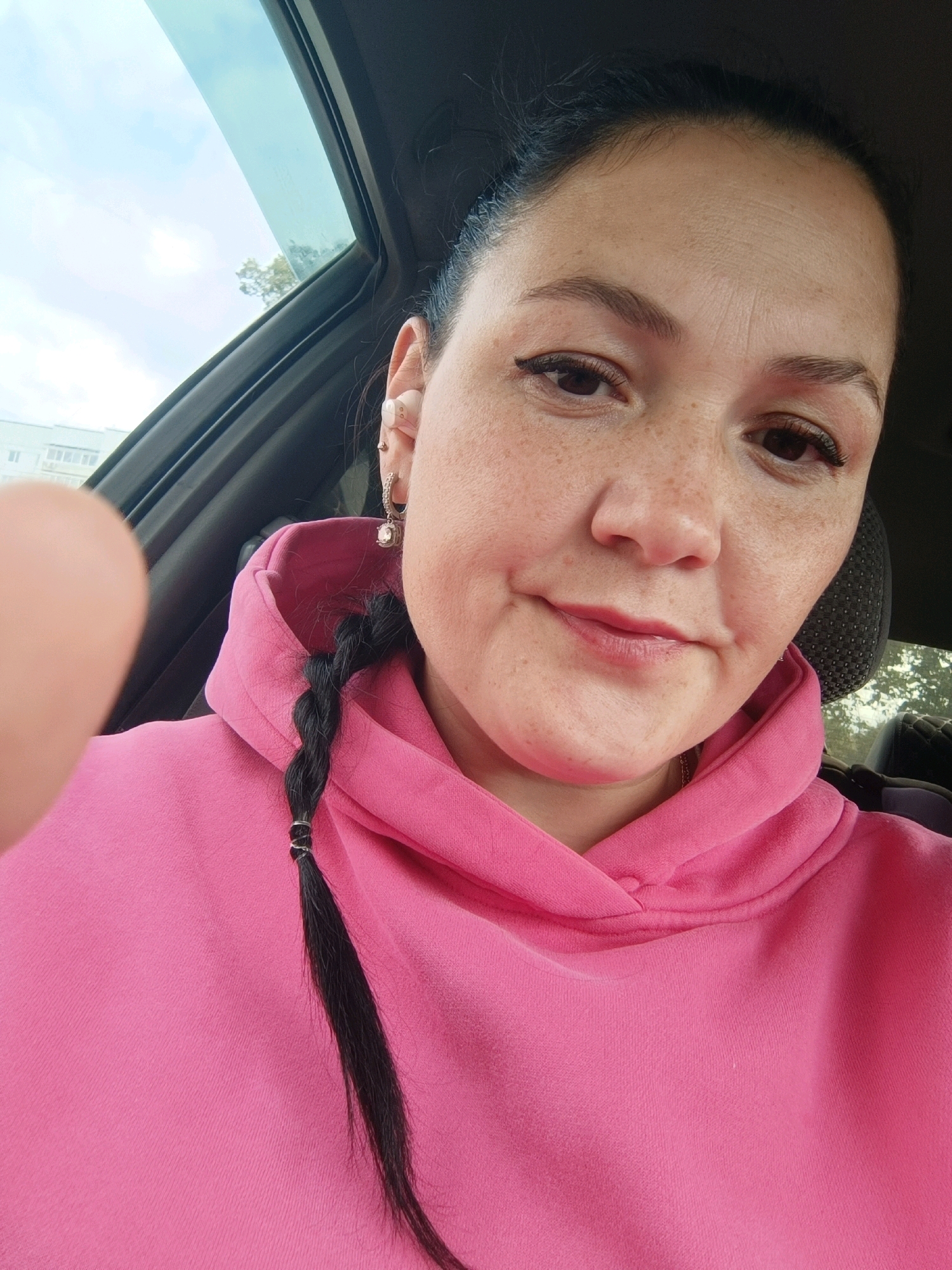 A woman with dark hair in a braid, wearing a pink hoodie, is inside a car. She has freckles on her face and is wearing an earring and an earbud. The window of the car is open, showing the sky and some trees outside. In the background, a residential building can be seen. The scene appears to be located in the city of Chaikovsky, Russia.