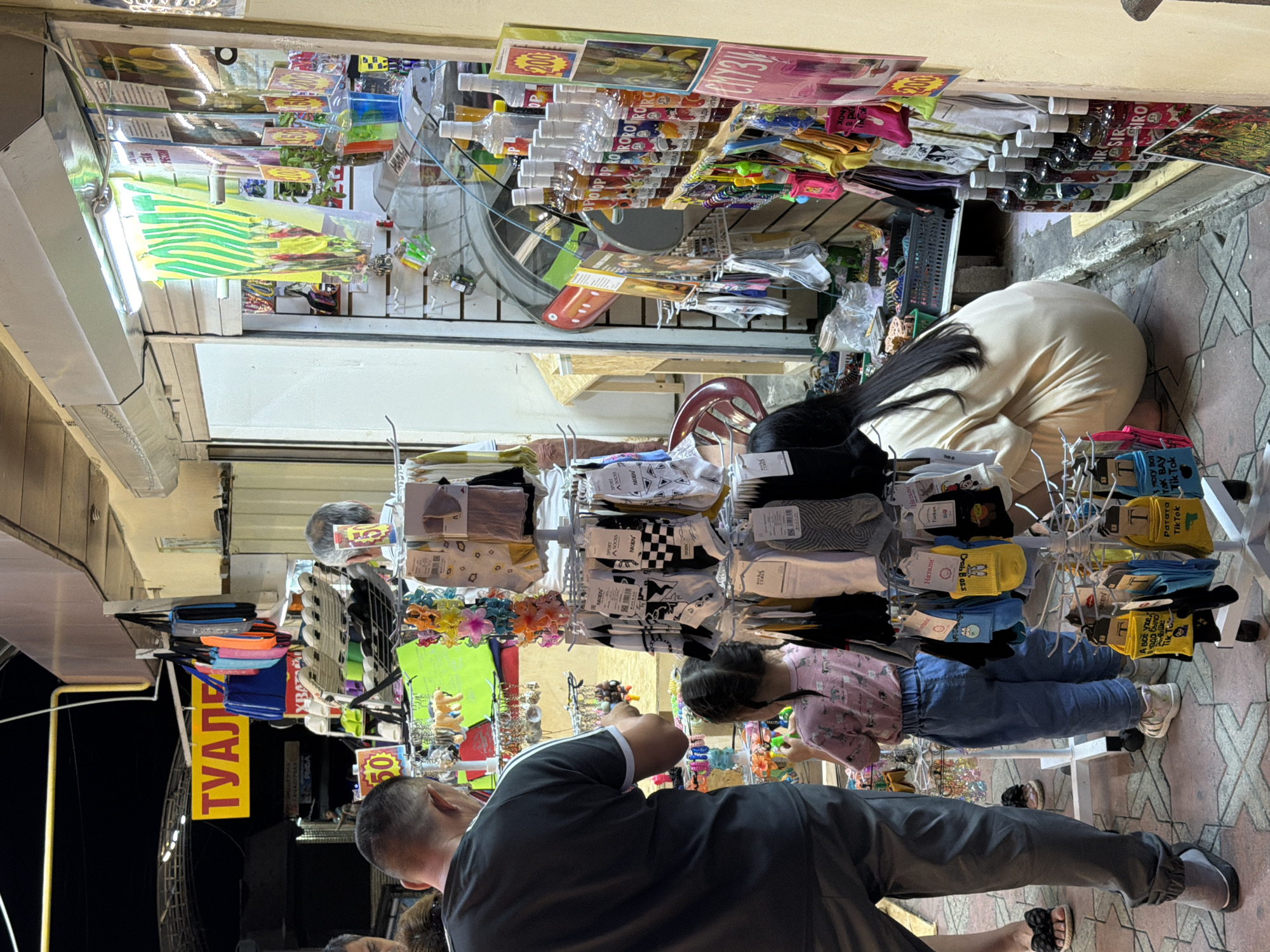 The image depicts a street market stall in Sochi, Russia, at night. In the foreground, a man in a gray t-shirt and pants is looking at merchandise. To his right, a young girl in a pink t-shirt and blue jeans is also examining items. Behind them, a woman in a light-colored dress is crouching down. The stall displays a variety of goods, including socks, bags, and bottles of what appear to be syrups or beverages. A prominent yellow sign with red lettering reads "ТУАЛЕТ" (Toilet). The stall is illuminated by fluorescent lights, and the ground is paved with patterned tiles. Other stalls and structures are visible in the background.