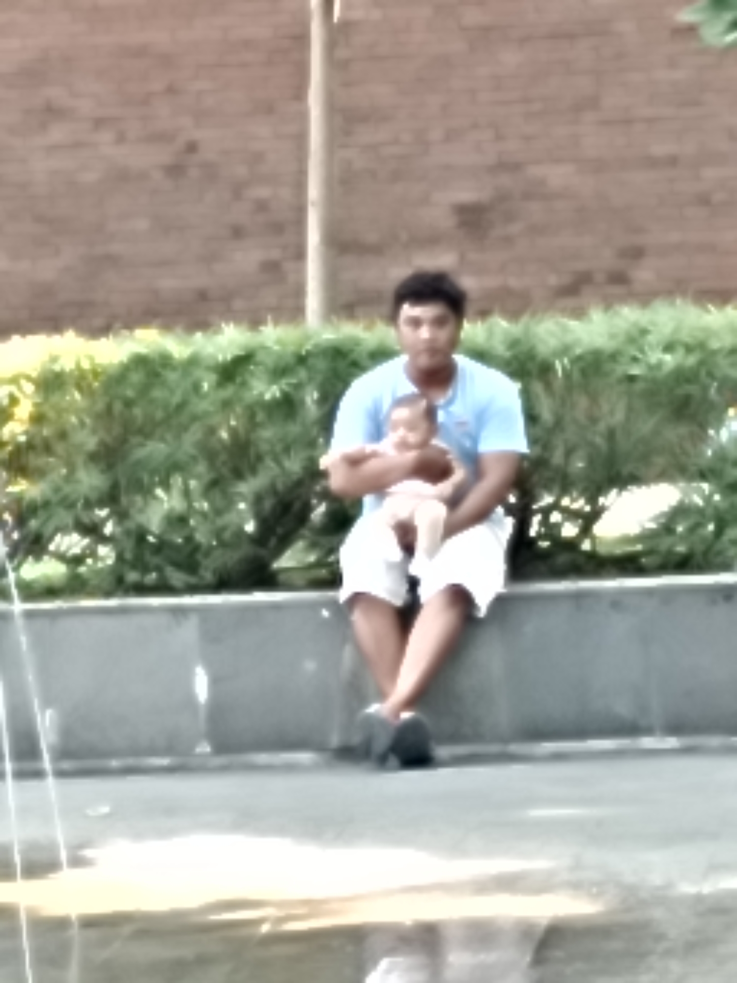 A person is sitting on a ledge outside, holding a baby. The person is wearing a blue t-shirt and white shorts. The baby is wearing a white outfit. They are sitting in front of a hedge with a brick wall behind it. The area appears to be a park or public space.