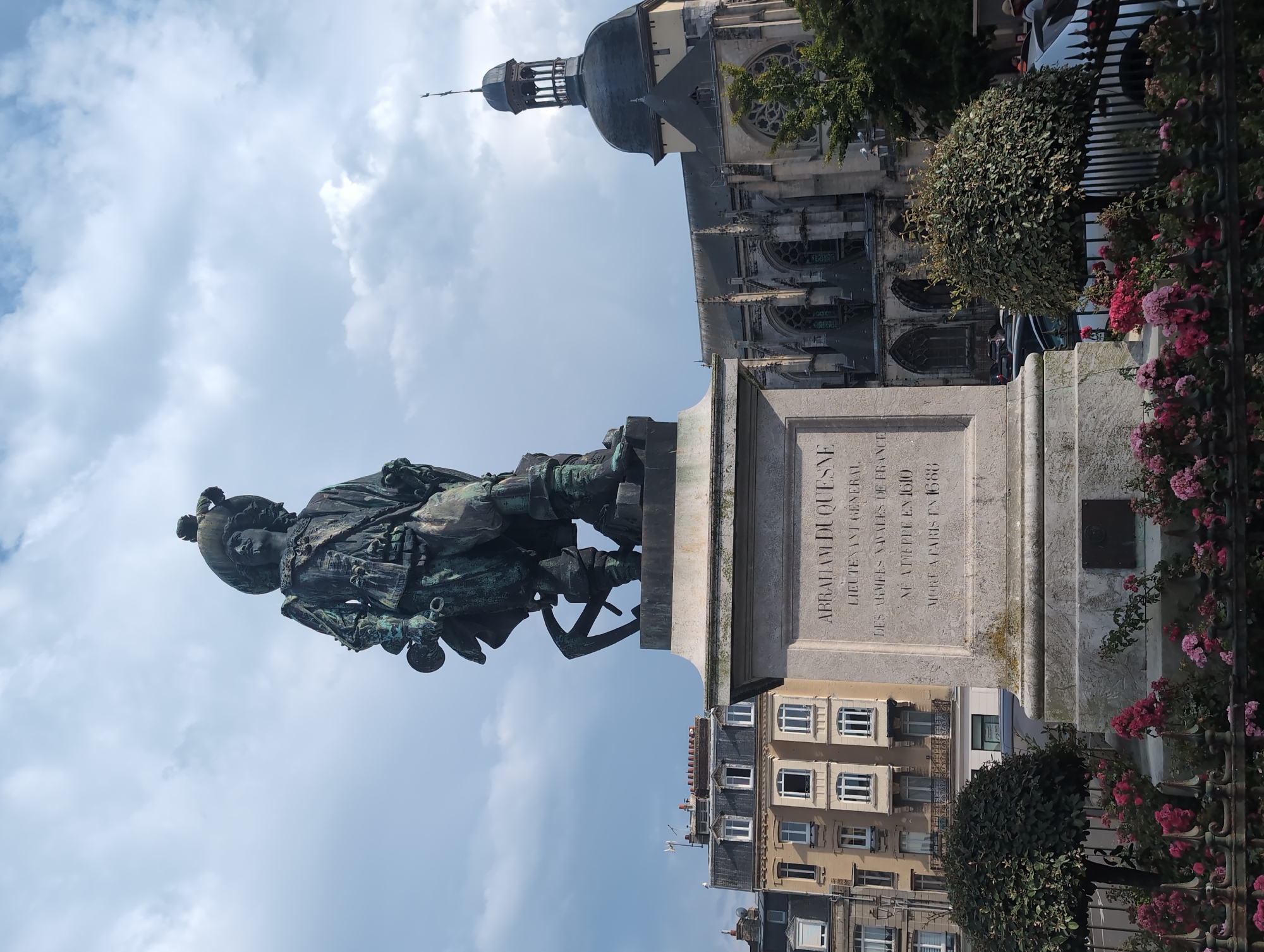 An eye-level, full shot captures a bronze statue of Abraham Duquesne in Dieppe, France. The statue depicts Duquesne, a lieutenant-general of the French navy, dressed in period attire, holding a spyglass in his right hand and a sword hilt in his left. He wears a feathered hat and knee-high boots. The statue stands atop a stone pedestal inscribed with "ABRAHAM DUQUESNE LIEUTENANT GENERAL DES ARMÉES NAVALES DE FRANCE NE À DIEPPE EN 1610 MORT À PARIS EN 1688". The pedestal is surrounded by a fence and a bed of pink roses.
In the background, to the left of the statue, is a building with several floors, featuring a beige facade and numerous windows with white shutters. To the right of the statue is the gothic architecture of the Church of Notre-Dame de Bonsecours, with its large arched windows and a prominent dome topped by a cross. The sky above is partly cloudy. The scene suggests a historical and civic commemoration in a public space.