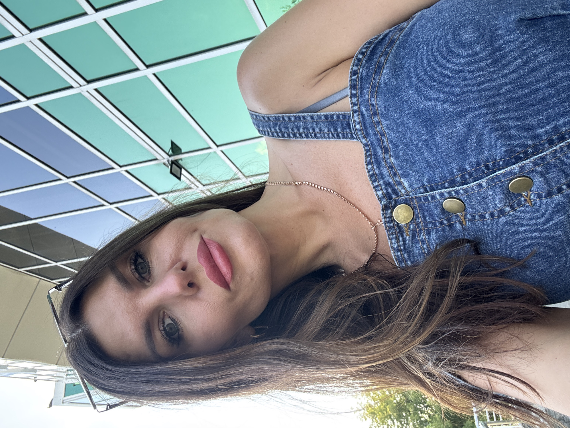 The media file is a close-up, portrait-style selfie of a young woman outdoors. She is wearing denim overalls over a light-colored top and a delicate rose gold necklace. Her dark brown hair is long and flows around her face and shoulders. Her makeup includes bold lipstick in a muted pink shade and dark eyeliner that accentuates her green eyes. She is wearing thin-rimmed glasses that rest on her cheek.
The background suggests an urban setting with a modern building featuring a facade of rectangular panels in varying shades of green and blue, reflecting sunlight. The lighting indicates it is daytime, likely sunny, given the bright reflections. The composition is tightly cropped on the woman's face and upper body, with the background providing a colorful and geometric contrast. No text is visible in the image. The overall impression is casual and self-assured. The location context of Chaikovsky, Russia, is noted but not visually apparent within the image itself.