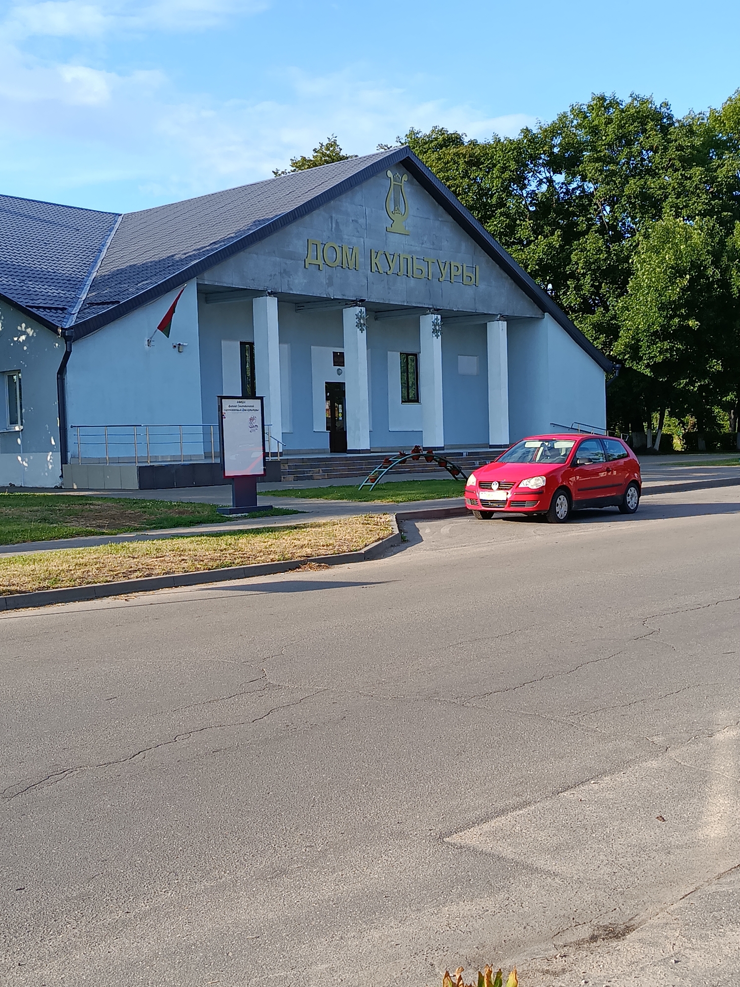 A bright red Volkswagen Polo is parked on the side of an asphalt road in front of a light blue building with a dark roof. The building has white columns and the text "ДОМ КУЛЬТУРЫ" written in gold above the entrance, along with a gold lyre emblem. A flag, red and green, is mounted on the building's facade. To the right of the building are tall green trees. In the foreground, the road shows cracks and wear. A sidewalk and a grassy area are between the road and the building. A digital sign on a dark stand is located to the left of the building's entrance.