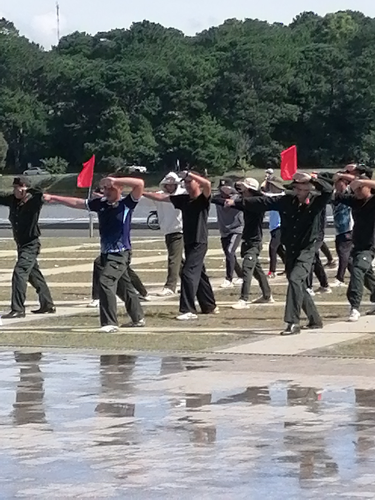 In Da Lat, Vietnam, a group of men in uniform are engaged in an outdoor exercise session on a paved area, possibly a sports field or public space, with a reflection of the scene visible on the wet ground in the foreground. The men are positioned in a line and appear to be performing synchronized movements, with several of them raising their arms and gesturing with their hands. They are dressed in dark uniforms, including shirts and pants, and some are wearing hats. Red flags are visible in the background, suggesting a possible organized event or training. A line of trees forms the backdrop, and a cloudy sky is overhead. The environment suggests an urban or semi-urban setting, given the paved surfaces and the presence of what appear to be traffic barriers and a distant building.