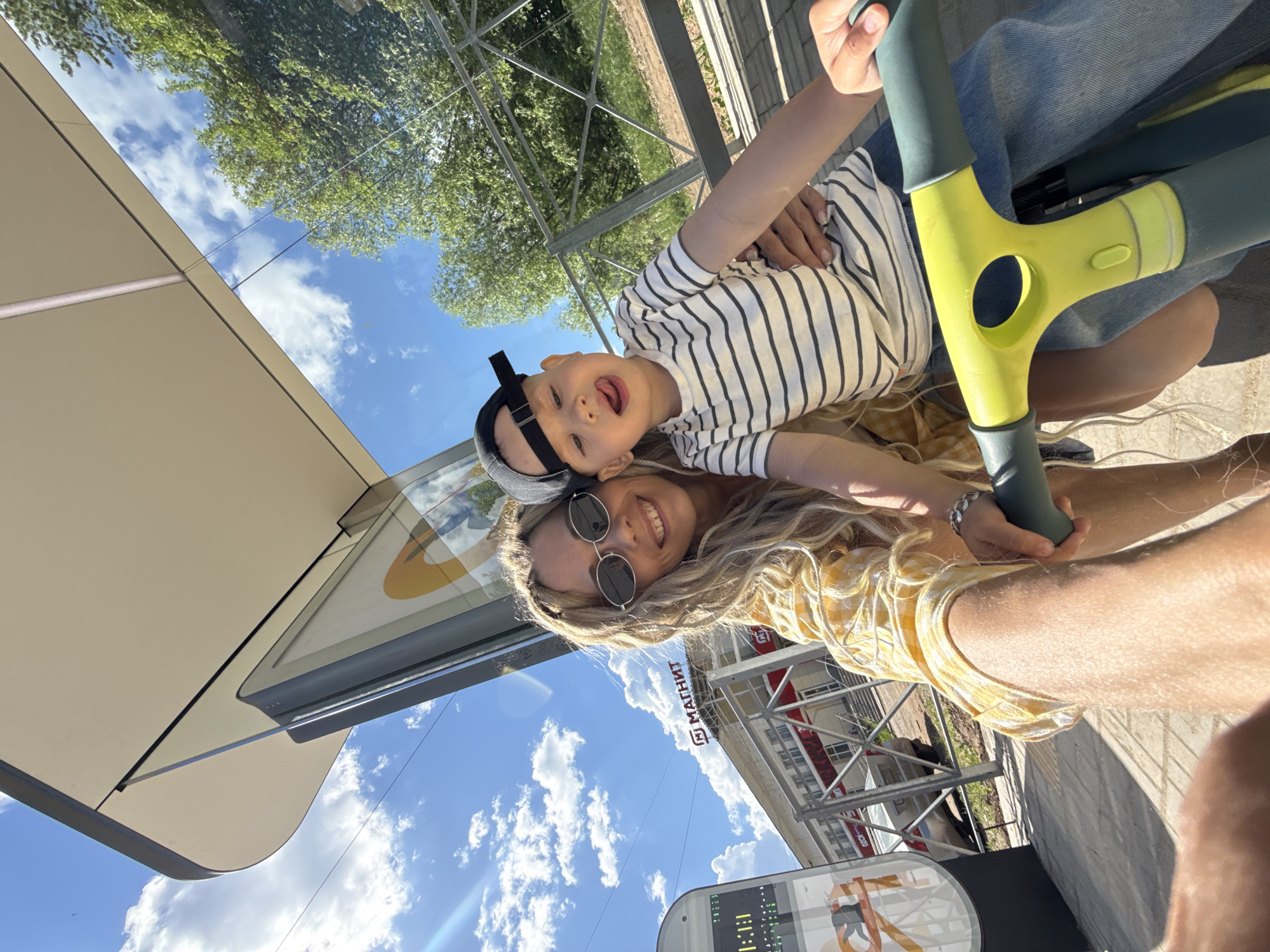 A woman wearing sunglasses and a yellow plaid shirt is holding a child wearing a black and white striped shirt and baseball cap. They are next to a light yellow and green scooter. In the background, there's a building with a sign that says "МАГНИТ", which indicates that this photo might be taken in the city of Rusanovo, Russia. The sky with clouds and some trees are visible.