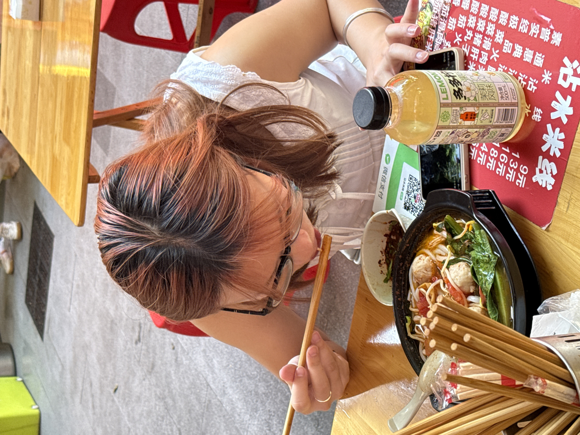 Here's a detailed description of the image:
The image shows an overhead view of a woman with dark hair, highlighted with reddish-brown sections, eating at a table. She's wearing glasses and a white top. She is holding chopsticks and appears to be eating noodles from a black bowl. The bowl contains noodles, meatballs, tomatoes, and green vegetables.
To the right of the bowl is a cup filled with unused chopsticks. A bottle of Lemon tea drink, with Chinese writing on the label, is also visible on the table. A smartphone can be seen next to the bottle.
A red menu card is present with Chinese characters, including the characters "沾水米线" (zhan shui mi xian) which translates to "dipping rice noodles." The menu lists prices for different varieties of the dish. Below that is a QR code labelled "微信支付" (wēi xìn zhī fù) which means "WeChat Pay".
The table is wooden, and a red chair is partially visible. The background suggests an outdoor dining area, with a gray pavement visible. The scene appears to be a local eatery in the city of Beibei, China, based on the language on the menu and the type of food.