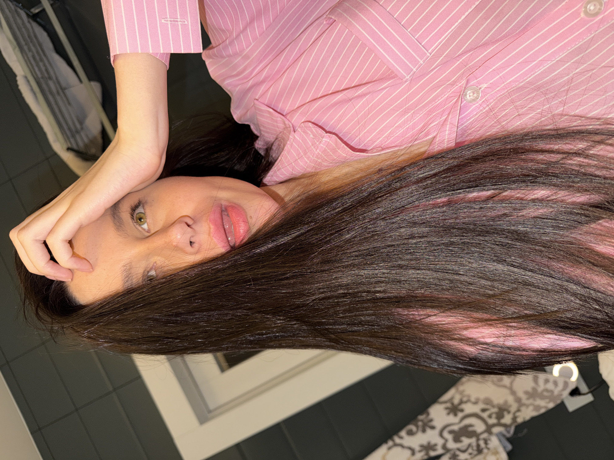 The primary image, presented sideways, features a person, likely a woman, lying down with her head tilted. Her long, dark brown hair spreads out, partially obscuring her face. Her left eye, which appears green or hazel, is visible and gazes directly forward. Her lips are slightly parted. She is dressed in a pink and white striped button-down shirt; the cuff distinctly shows the brand name "RALPH LAUREN" in subtle text. Her right arm is bent at the elbow, with her hand resting on her forehead, revealing part of her smooth forearm.
The setting is an indoor space, characterized by a dark, possibly tiled, floor in the foreground and a white, angular architectural element in the mid-ground, which could be a window frame or furniture. A patterned fabric, possibly a rug or cushion with a light-colored floral or paisley design, is visible in the bottom right corner. The lighting suggests an indoor, likely artificial, light source, contributing to a calm and somewhat intimate atmosphere. The person's expression is neutral to slightly pensive. The overall scene suggests a casual, perhaps relaxed moment, in a contemporary interior, located in Khimki, Russia.