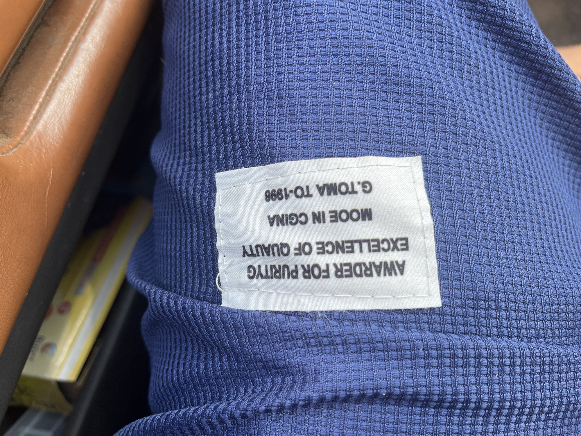 The image shows a close-up of a blue, textured fabric with a white label attached. The label has black text printed on it. The text reads: "AWARDER FOR PURITYG EXCELLENCE OF QUAUTY MOOE IN CGINA G.TOMA TO-1998". The fabric appears to be a garment, possibly a shirt or jacket. The background is blurred, but suggests an indoor setting with some objects in the distance, possibly a brown leather surface and a yellow item with some writing. There is no visible indication of the city of Aqköl, Kazakhstan, or any specific location within it.