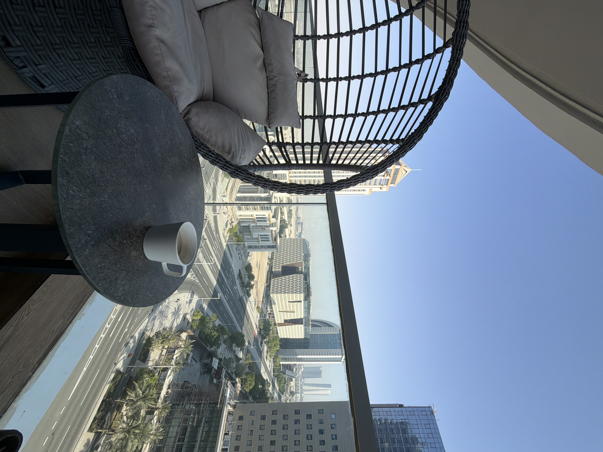 An outdoor balcony offers a view of a modern cityscape under a clear blue sky. A round, dark gray textured table sits in the foreground, holding a white mug of coffee. To the left of the table is a large, dark gray rattan hanging chair with light-colored cushions. Beyond the balcony railing, a street with multiple lanes of traffic runs through the city, flanked by various architectural structures, including contemporary buildings and a distinctive building with a patterned facade. Palm trees and other greenery line the street. The scene is captured in daylight, with soft shadows indicating the time of day.