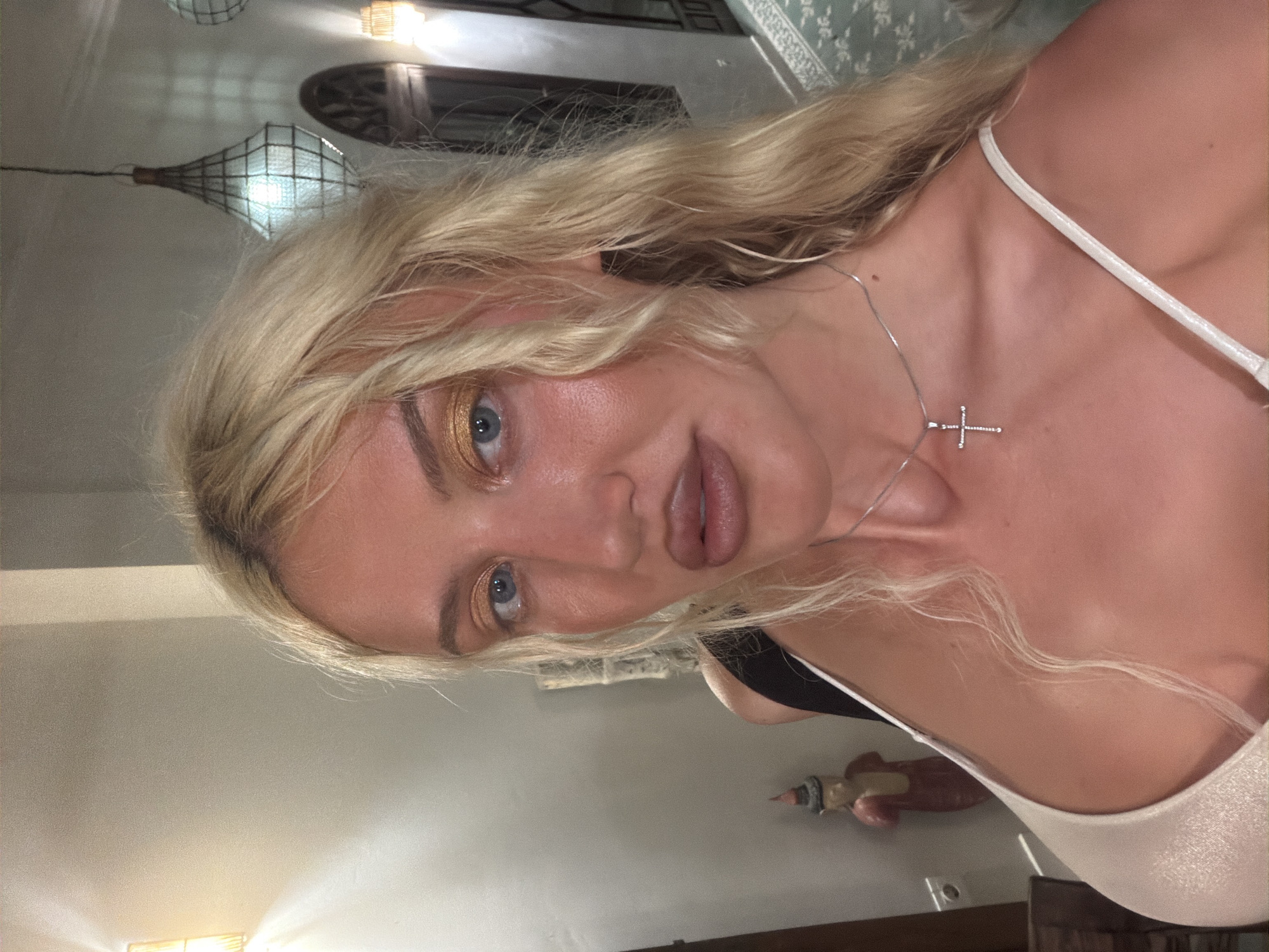 A woman with blonde hair and blue eyes is taking a selfie. She is wearing a sparkly white top and a silver necklace with a cross pendant. The background appears to be a hallway or room with decorative lamps and artwork. The scene is likely indoors, potentially in a hotel or residence in North Kuta, Indonesia.