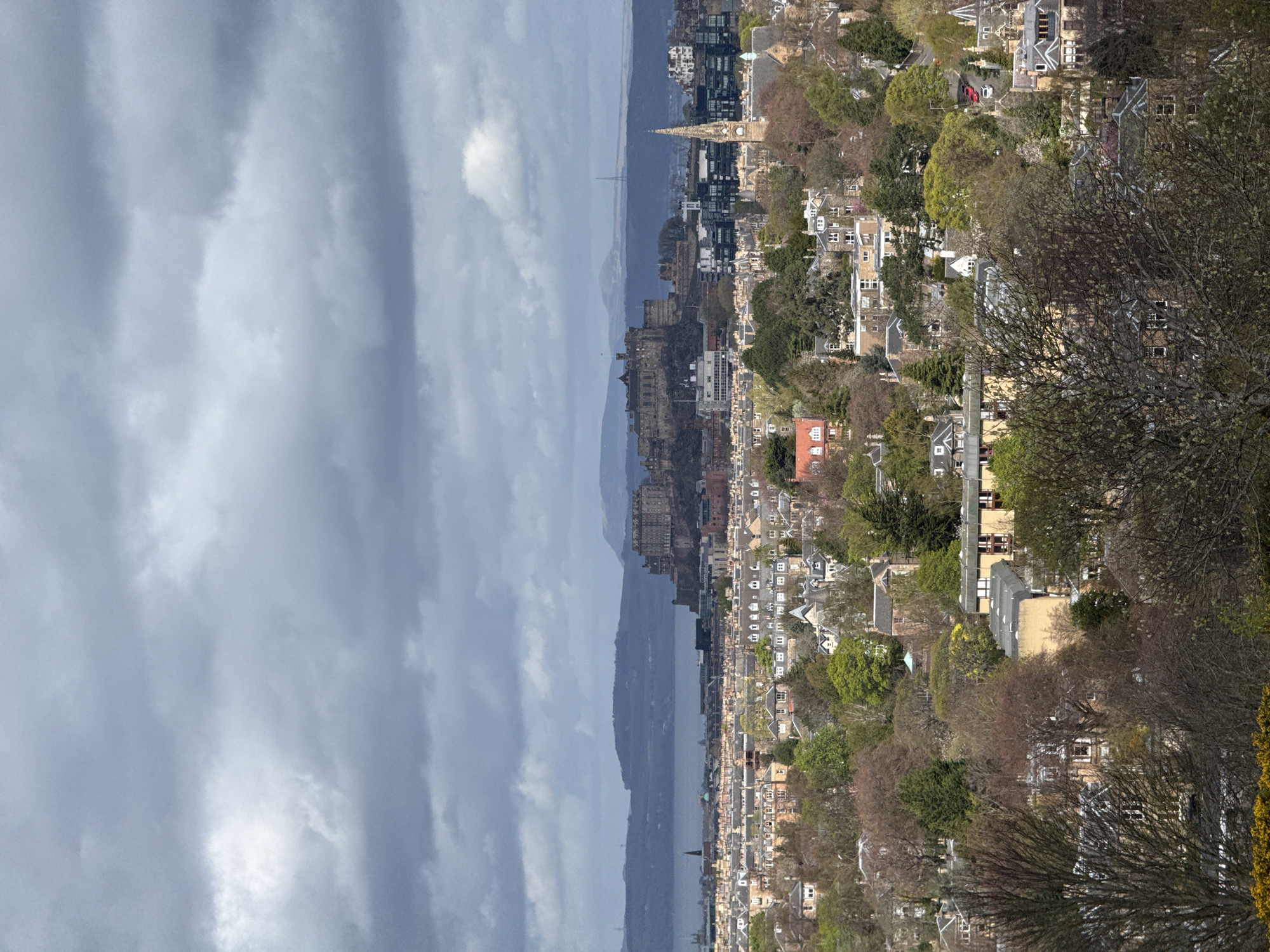 This panoramic image, rotated 90 degrees clockwise, presents a wide view of a city and its surroundings under an overcast sky. The overall scene is dominated by a sprawling urban landscape extending towards a large body of water, with distant hills fading into the horizon.
The sky is filled with thick, grey clouds, indicating an overcast day, though some lighter patches suggest indirect sunlight might be breaking through. In the foreground, on the right side of the correctly oriented image, a dense residential area is visible, with numerous buildings nestled amongst a mixture of bare and leafy trees, suggesting either early spring or late autumn.
The midground features a prominent body of water, likely a firth or bay. Overlooking this water, a large, historic-looking castle or fortified structure stands on a rocky outcrop, dominating the skyline. To the right of the castle, a significant part of the cityscape unfolds, characterized by a mix of older and more modern buildings, including a church spire rising above the rooftops. Faintly visible in the far distance, across the body of water, is a bridge structure.
The background consists of a long stretch of hills or mountains on the opposite side of the water, appearing hazy and blending with the muted sky. No specific activities or people are discernible. The light suggests it is daytime, and the cloudy weather creates a somewhat muted and atmospheric mood. There is no visible text in the image. The prominent castle overlooking a firth, alongside a large city, strongly suggests the location is Edinburgh, Scotland, with the castle being Edinburgh Castle and the water being the Firth of Forth.