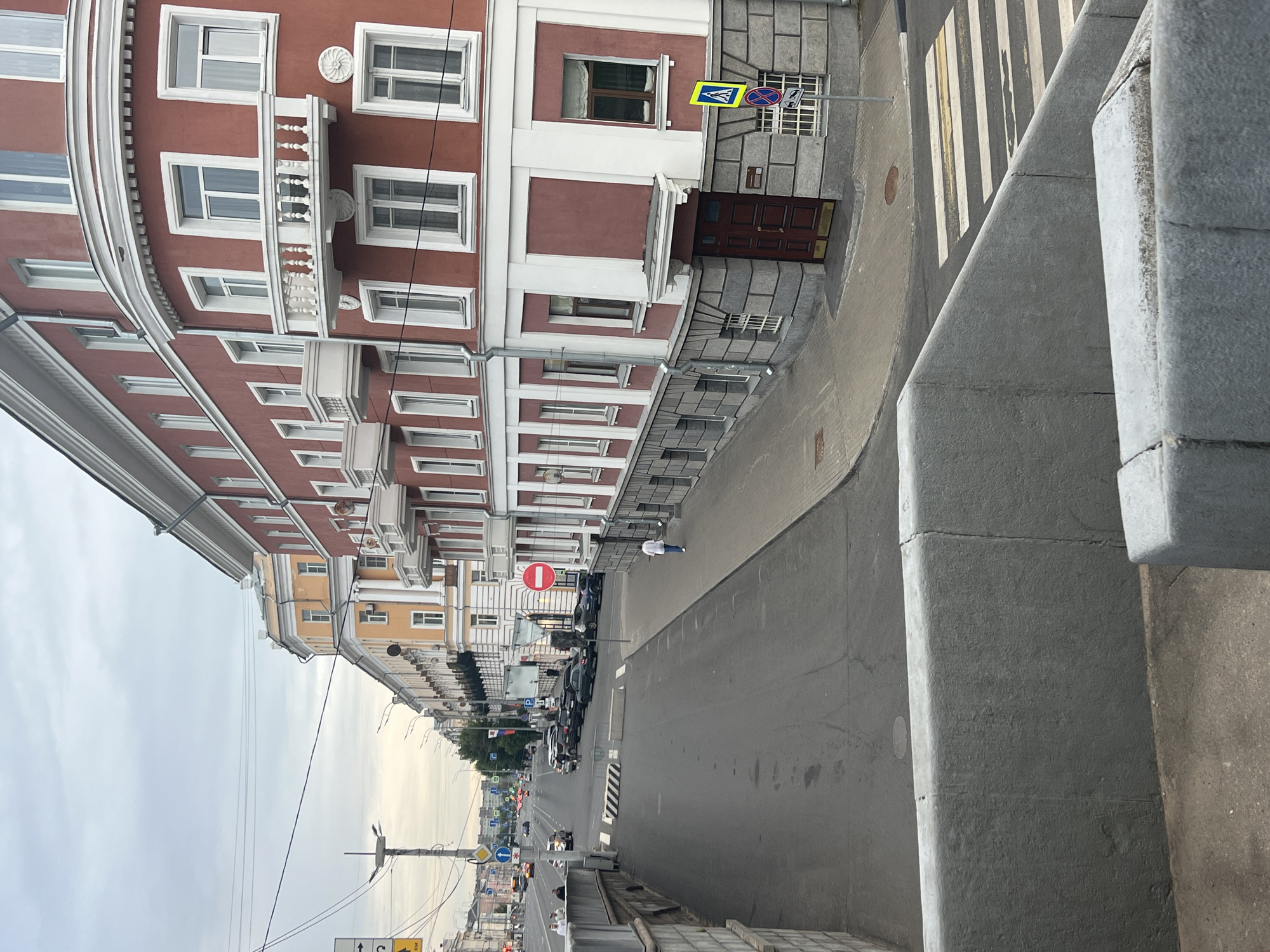 The image shows a street scene in Tver, Russia, likely in the city center, given the architecture and urban environment. The view is from an elevated position, looking down a wide, paved street lined with multi-story buildings. On the right, a prominent red building with white trim and balconies stands out. It has numerous windows and a classical architectural style. To the left, other buildings with lighter facades, also multi-story, continue down the street. Several cars are parked along the side of the street, and traffic is visible further down the road, including cars and potentially a bus. A pedestrian crossing is visible in the foreground on the right side of the street, marked by white and yellow stripes. A person in a white shirt and blue jeans is walking away from the camera on the sidewalk in front of the red building. Traffic signs, including a red circular "no entry" sign and a blue pedestrian crossing sign, are present. The sky is overcast, suggesting a cloudy day. The overall scene depicts a typical urban street in a Russian city.