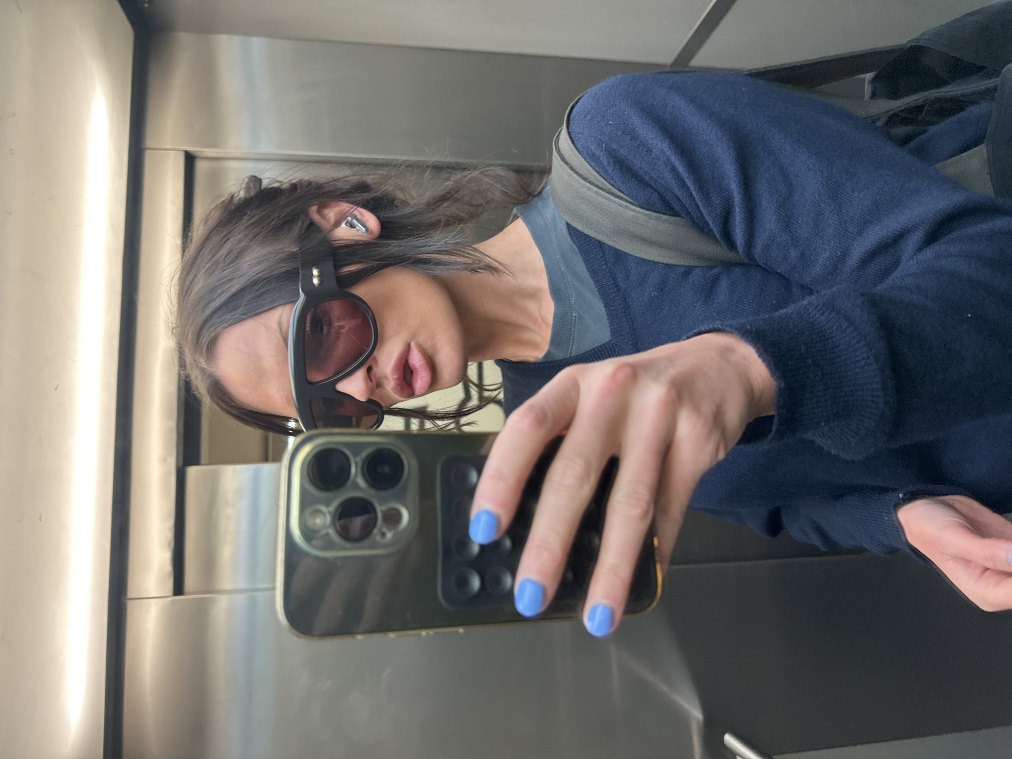 A person is captured in a selfie taken in a reflective metallic space, likely an elevator or a modern interior with polished walls, possibly in London, United Kingdom. The individual has long, dark hair and is wearing large, dark-rimmed sunglasses with tinted lenses. A silver-colored earbud is visible in their right ear.
They are dressed in a dark blue long-sleeved sweater over a dark grey t-shirt. A grey strap, suggesting a backpack, is visible over their left shoulder. The person is holding a smartphone, identifiable as an iPhone by its distinct multi-lens camera module on the back, encased in a clear cover. Their left hand, holding the phone, features light blue nail polish. The selfie captures their reflection, with lips slightly parted, creating a neutral expression partly obscured by the sunglasses. The background shows seams of the metallic panels and bright, artificial overhead lighting. No text is visible in the image.