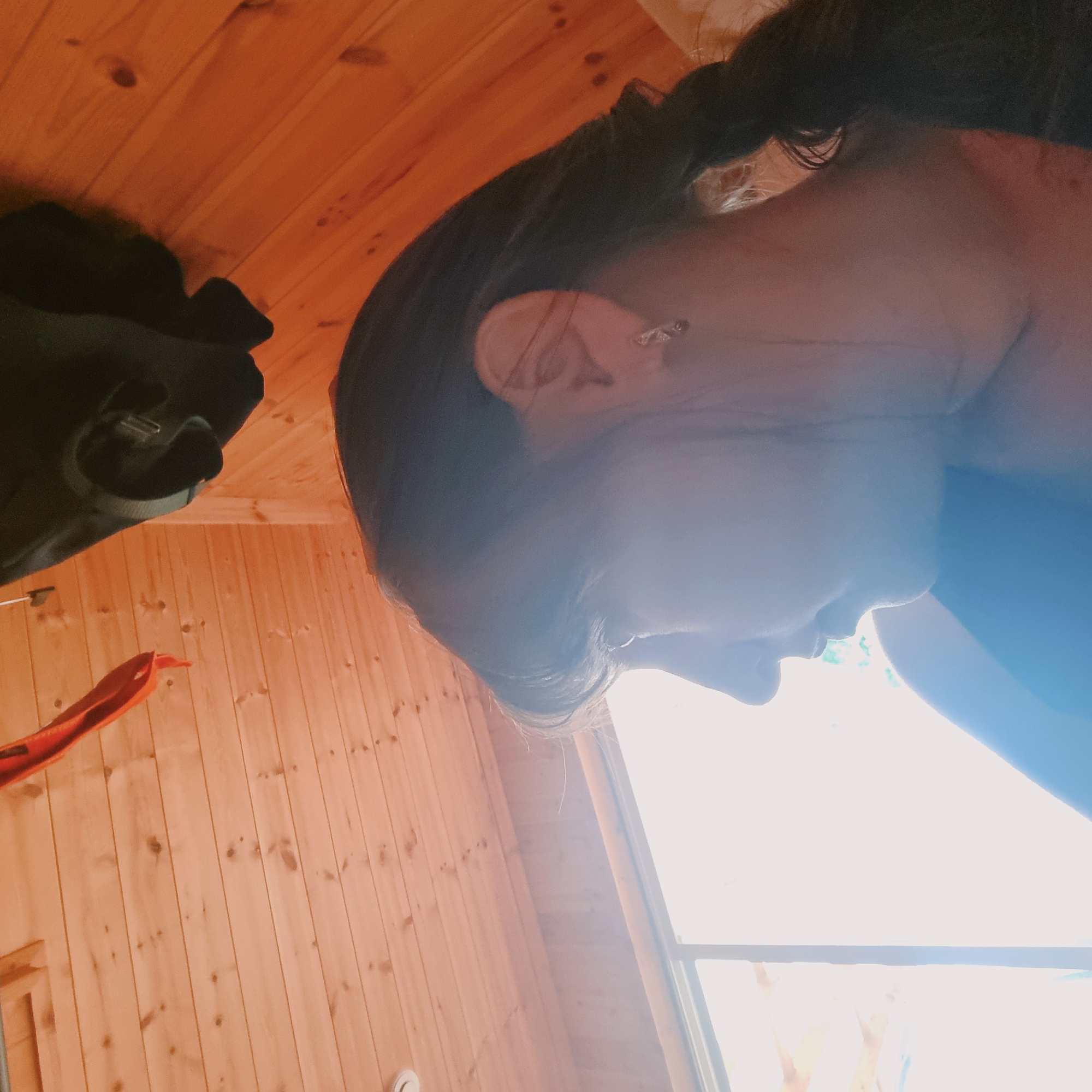 The image shows a woman from the side, looking out a window. She has dark hair pulled back in a braid and is wearing a small earring. The room has wooden walls and ceiling, and a bright light comes in from the window. There is a black bag hanging on the wall. The setting appears to be a rustic cabin or a room with a natural wood interior.