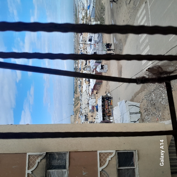 View from a window with a metal grate. Below is a building with two windows decorated in geometric patterns. Beyond the grate, a dense settlement of makeshift structures and tents stretches towards the sea. A road with white markings runs along the edge of the settlement. A blue sky with light clouds is visible above the settlement. The image orientation is vertical. "Galaxy A14" is printed in the bottom right.