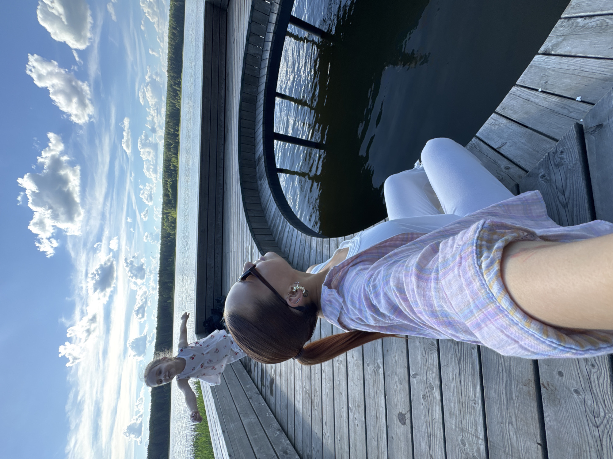 In Votkinsk, Russia, on a wooden pier extending over a body of water, a woman and a young girl are present. The woman is seated on the pier, facing away from the camera, with her head tilted upwards. She is wearing white pants, a white tank top, and a pink and purple plaid shirt. Her hair is pulled back in a red ponytail, and she has sunglasses on and earrings. The young girl is standing on the pier behind the woman, with her arms outstretched, looking towards the sky. She is wearing a white dress with red strawberry patterns. The pier is constructed of wooden planks and features a curved section with vertical supports extending into the dark water. In the background, a dense green forest lines the opposite shore of the water, under a bright blue sky with scattered white clouds. The scene appears to be outdoors in daylight.
