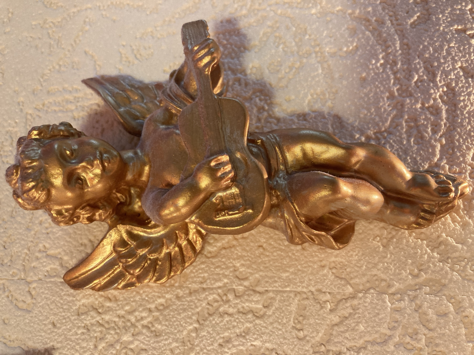 An ornate golden cherub figurine is attached to a textured, cream-colored wall. The cherub is depicted in a dynamic pose, leaning forward with wings outstretched, and is holding a small guitar. The figurine has a metallic sheen, reflecting the light. The wall exhibits a rough, stucco-like texture with subtle shadows and highlights that emphasize its uneven surface. The lighting suggests an indoor setting, possibly a domestic environment. The specific location in the city of Unknown, Unknown cannot be determined from the visual information provided.