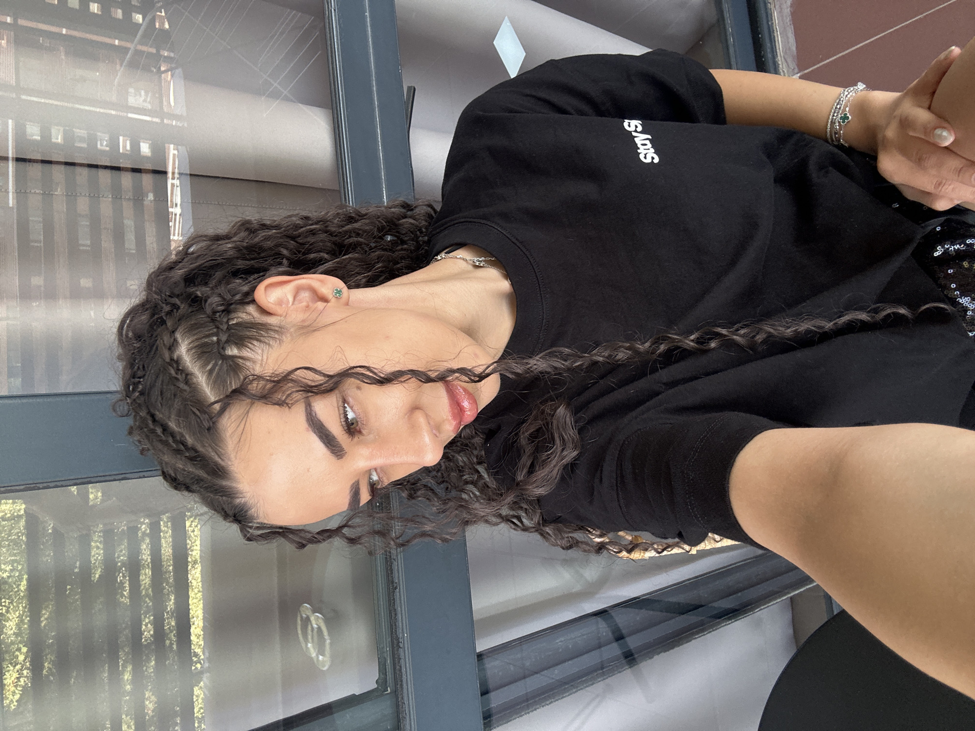 A woman with dark curly hair styled in braids and loose curls, wearing a black t-shirt with white text reading "Stay Si" is pictured in a close-up selfie. She has a green clover-shaped earring in her left ear and a silver necklace. She is wearing several silver bracelets on her left wrist, one of which also has a clover charm. She is looking off to her left, with her head tilted. Her left arm is visible, resting on her lap. In the background, there are large windows with horizontal blinds and sheer curtains, suggesting an indoor setting with natural light. The environment appears to be modern and possibly a hotel or apartment with a view, though no specific city landmarks are identifiable. It is not possible to determine the location within Nesebar, Bulgaria from the provided image.