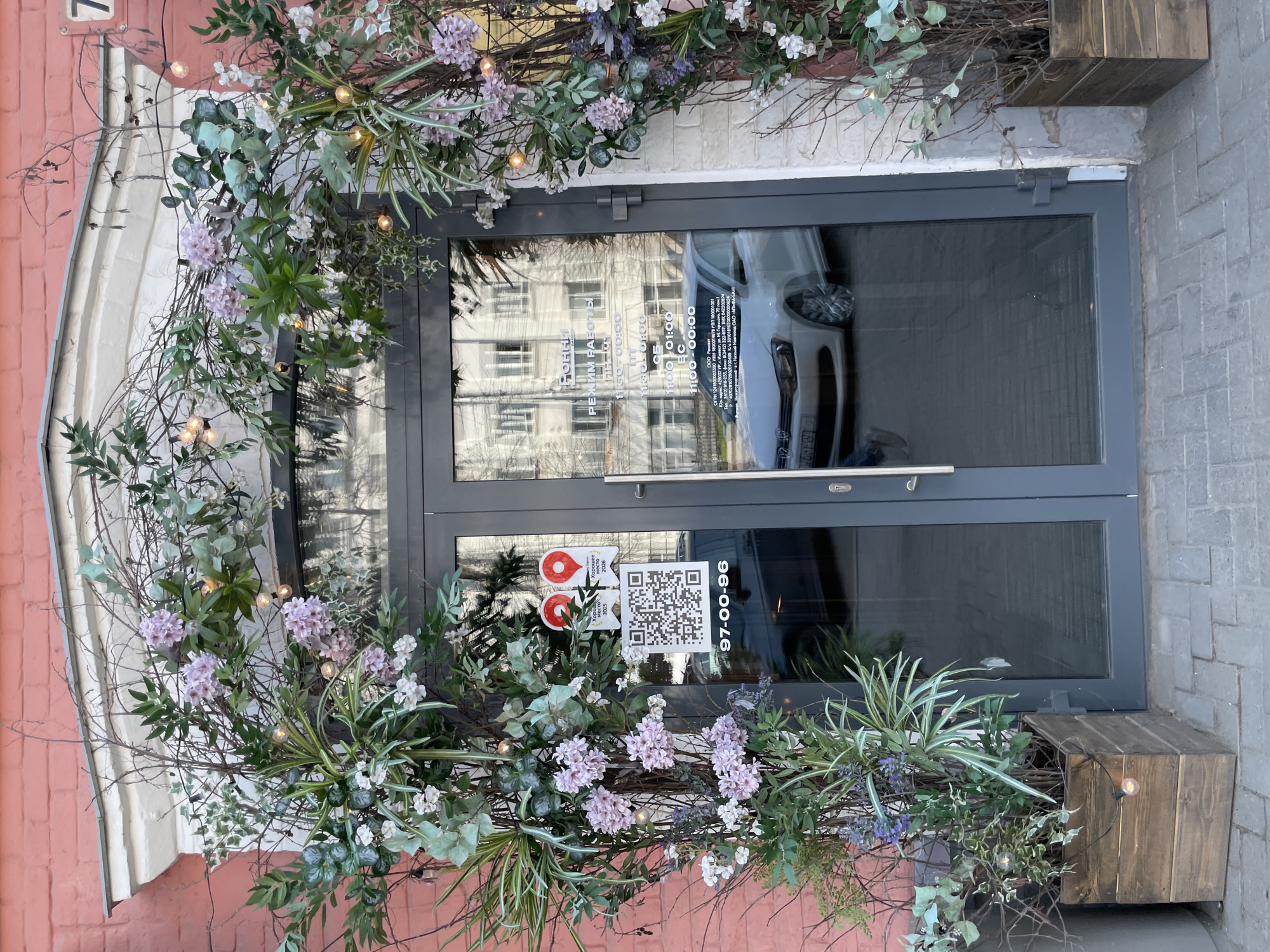 The image captures the entrance to a business establishment, likely a shop or restaurant, located at 7 M. Gorkogo Street in Izhevsk, Russia. The entrance features a glass double door with a gray frame, decorated with an arch of flowers, branches, and string lights.
A QR code and stickers are affixed to the glass, indicating the business is a "Good Place" and including a phone number, 97-00-96. The door has a window with reflected buildings and a car outside. The business hours are posted in Russian and are partially obscured. The information appears to be from "OOO Rassvet", including an address, tax ID, and bank information.
The business is on the ground floor of a pink brick building with white trim around the door. The entrance is flanked by wooden planter boxes also holding greenery and string lights. The sidewalk pavement is visible in the lower right corner.