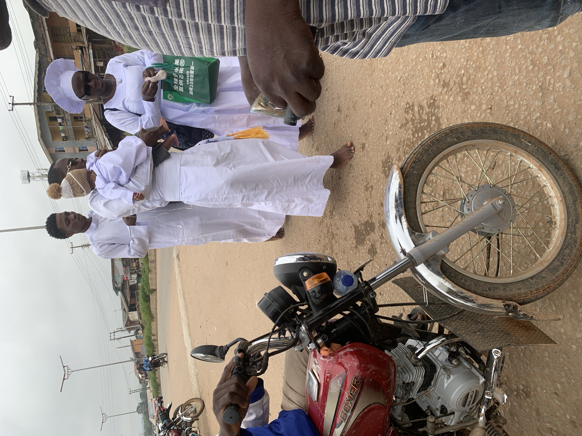 In Shagamu, Nigeria, a group of people in white robes stand on a dirt road. A person in the foreground is holding money, while another, wearing a striped shirt, appears to be taking it. A red motorcycle is parked to the left, and a woman in a white cap holds a green bag. The background shows buildings, streetlights, and a cloudy sky.