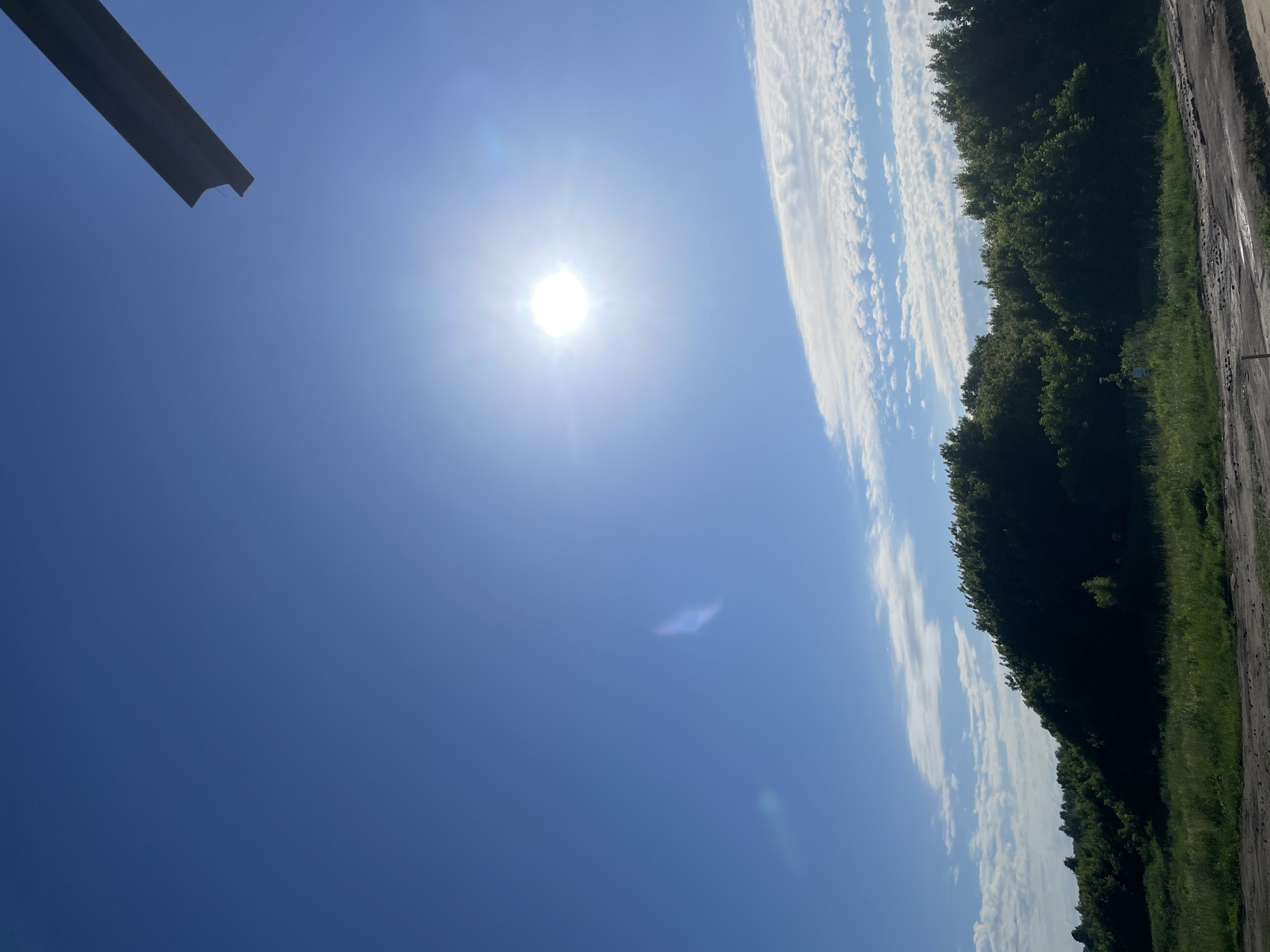 The image shows a bright, sunny day with a clear blue sky. The sun is high in the sky, casting a bright glare. A dark, angular object is visible in the upper right corner of the frame, possibly a piece of construction material or a piece of equipment. Below the sky is a dense line of green trees, forming a natural horizon. In the foreground, there is a patch of dry, dusty ground, likely a road or construction site, with scattered small rocks and debris. The overall scene suggests an outdoor, possibly rural or semi-constructed environment, consistent with the outskirts of a settlement like Сокуры, Russia, on a clear day.
