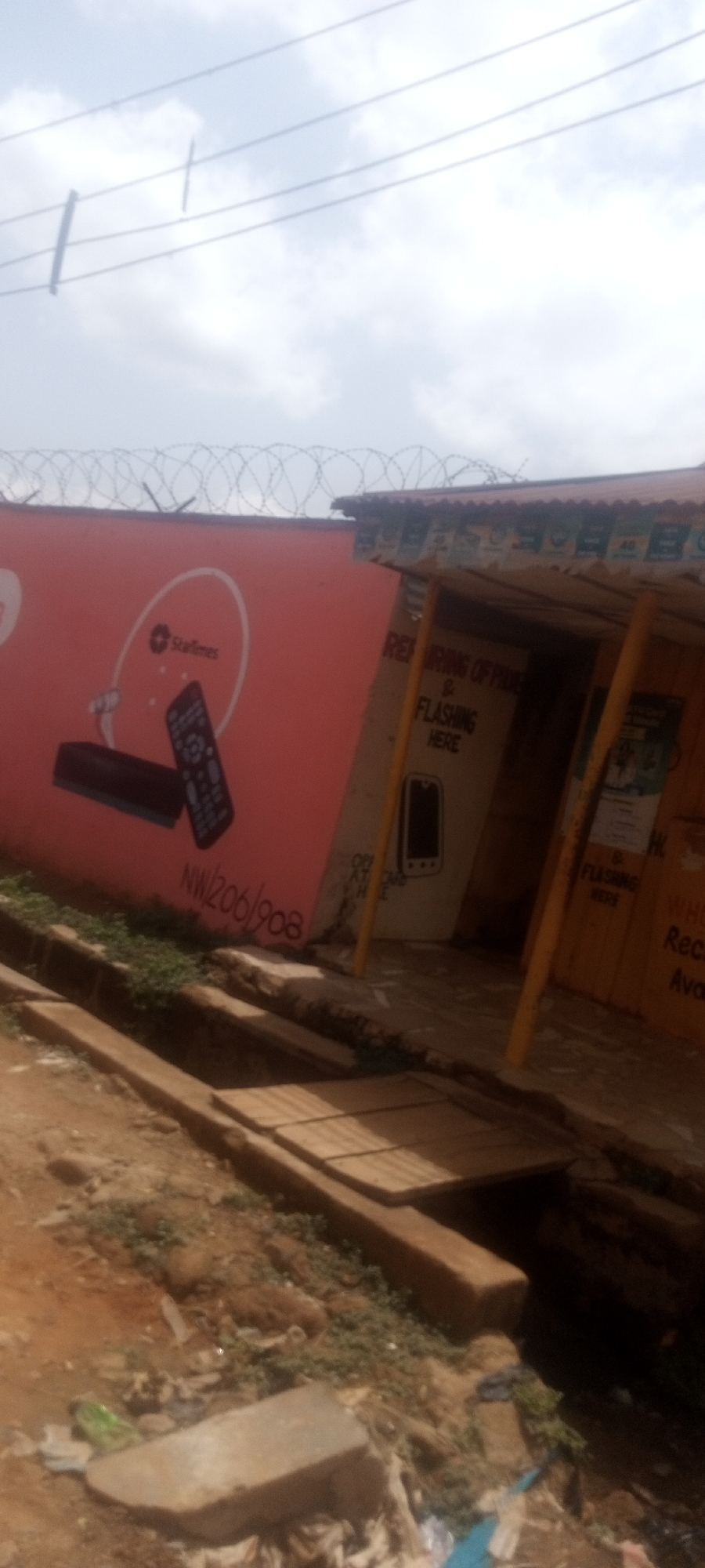 The image shows a shop located in Narayi, Nigeria. The shop, identified by its signage, offers phone repair and flashing services. The building adjacent to the shop is painted orange and features an advertisement for StarTimes. Barbed wire runs along the top of the wall. A ditch runs in front of the buildings, with a wooden plank bridging it. Litter is scattered in the foreground. Overhead power lines stretch across the cloudy sky.