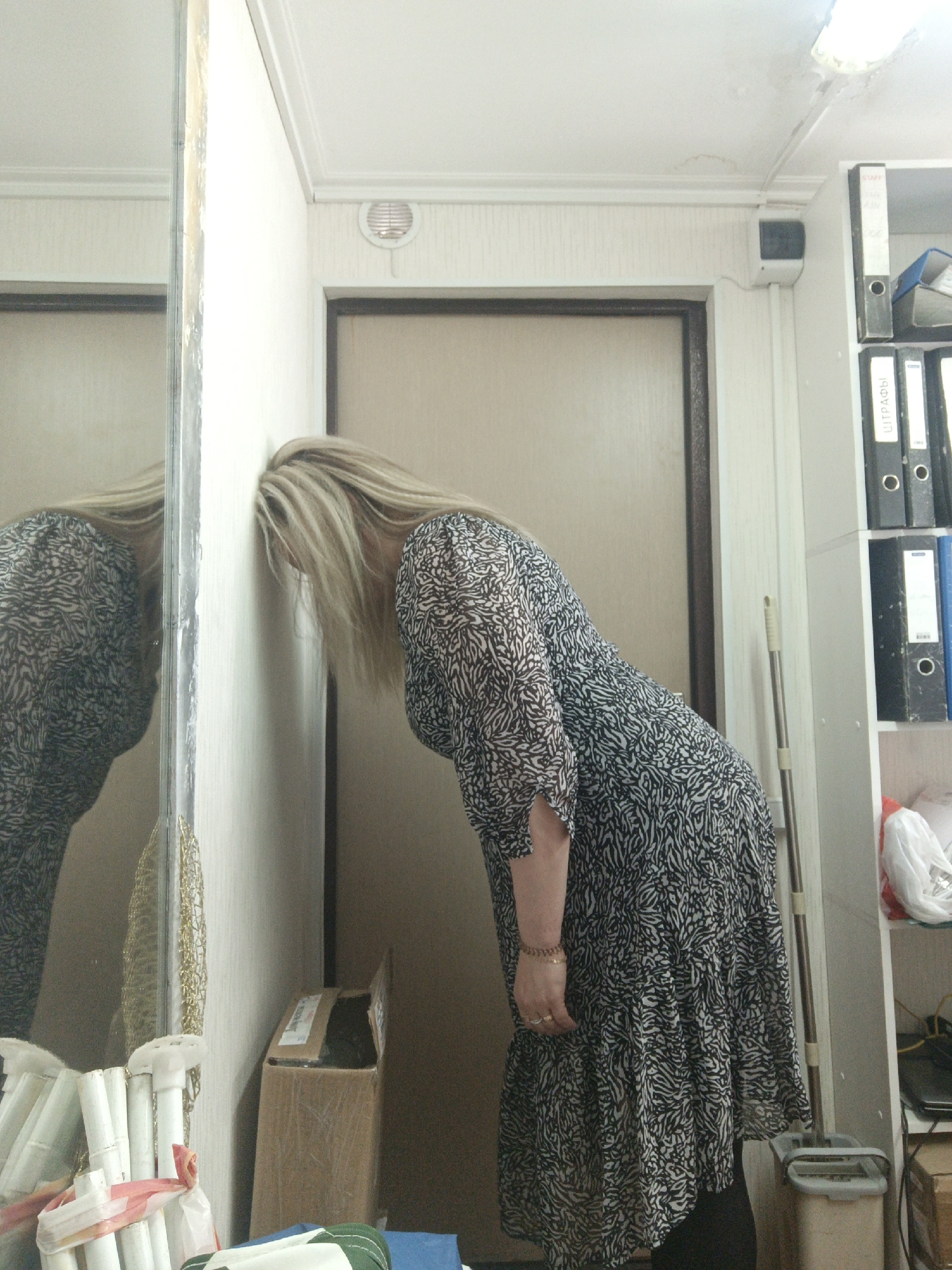 The image displays an indoor scene, likely located in Noviy, Russia, focusing on a person leaning against a white wall with their face obscured by their long, blonde hair. The person is wearing a black and white patterned dress, and their reflection is visible in a mirror that runs along the left side of the frame.
The posture of the person, with their head pressed into the wall, conveys a sense of frustration, exhaustion, or feeling overwhelmed. The setting appears to be a functional room, possibly an office or storage space. To the right of the person, a white shelving unit is stocked with several black binders. Visible text on one binder's spine reads "ШТРАФЫ" (SHTRAFY), which translates to "FINES" or "PENALTIES" in Russian. Another binder displays "STAFF" and numerical information.
Notable details of the environment include a partial view of a brown door frame behind the subject. A round ventilation grate is affixed to the wall near the ceiling. The ceiling itself features a fluorescent light fixture with visible damage and what appear to be water stains or marks. On the floor, a cleaning tool, possibly a mop or broom, leans against the shelving unit next to a beige bin or bucket. Near the bottom of the mirror, several white cylindrical objects are bundled together, and a golden, mesh-like decorative item is also visible. A cardboard box rests on the floor in the foreground. The person's right hand, adorned with a bracelet and rings, is partially visible.