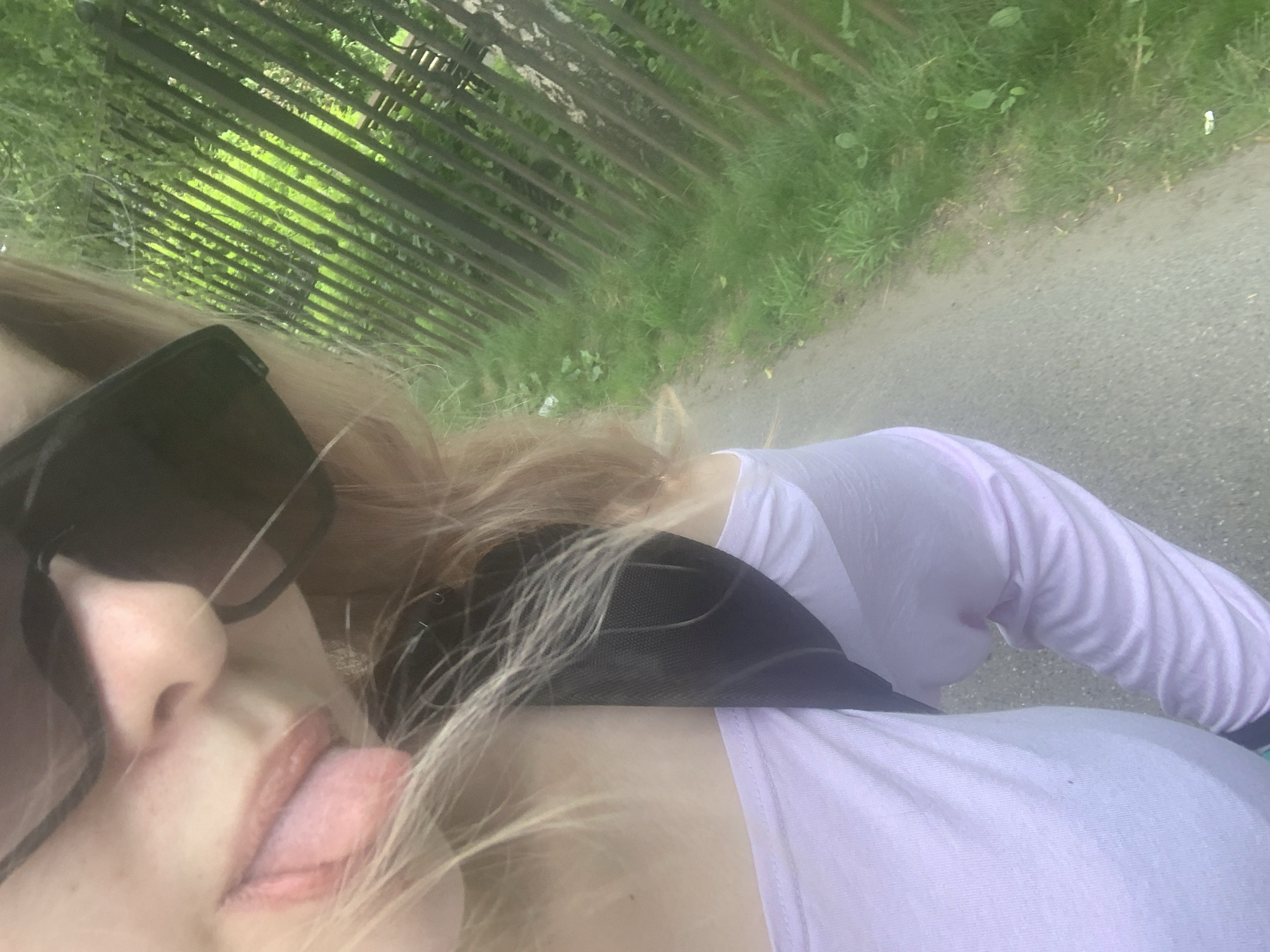 The image displays a close-up, first-person perspective of a person outdoors in the city of Unknown, Russia. The subject, a person with blonde hair and light-toned skin, is visible from the chest upwards. They are wearing dark sunglasses that obscure their eyes, and their tongue is slightly extended from their mouth. They wear a light purple long-sleeved top, with a black strap (likely from a backpack or bag) visible over their right shoulder. To the right of the subject, a dark metal fence with vertical bars separates a paved path from a dense green area featuring grass and foliage. The paved path extends along the lower left and bottom portions of the image.