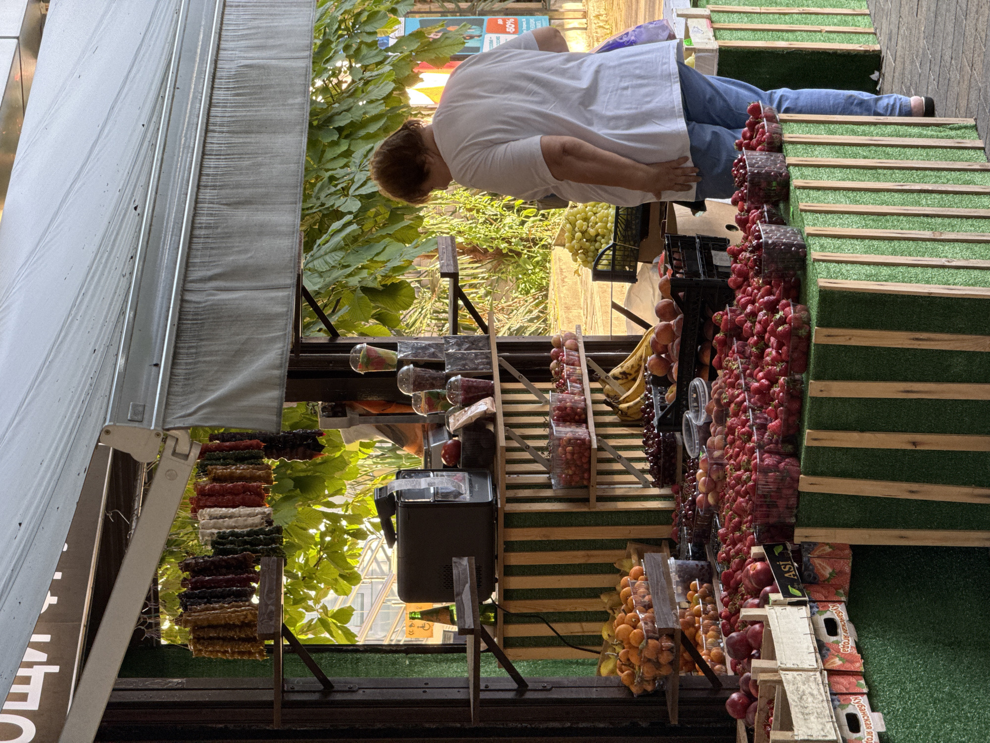 A woman wearing a white t-shirt and blue jeans stands at a fruit stand in Sochi, Russia. The fruit stand is outdoors, with a white awning overhead. The stand is filled with various fruits, including strawberries, cherries, apricots, bananas, and grapes. The woman is looking at the fruit. There are also some dried fruit snacks hanging from a rack above the fruit. In the background, there are trees and buildings.