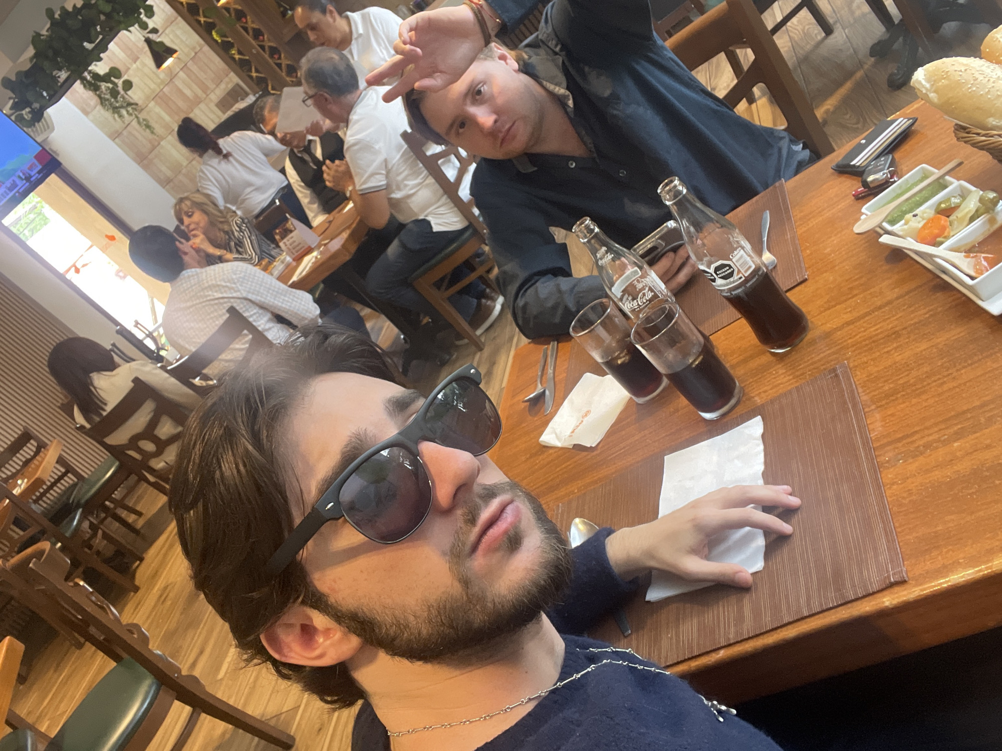 The image depicts a group of people dining at a restaurant. In the foreground, a young man with dark hair and a beard, wearing sunglasses and a dark blue sweater, is taking a selfie. He is seated at a wooden table with a brown placemat. Next to him, another man in a dark shirt is visible, making a peace sign with his hand. On the table, there are Coca-Cola bottles, glasses, napkins, and a small dish with what appears to be food. In the background, other diners are seated at tables. The restaurant appears to have a rustic interior with wooden furniture and decorative elements on the walls. The presence of Coca-Cola and the overall ambiance suggest a casual dining establishment. Based on similar architectural and interior design elements found in images of restaurants in Cuautitlán Izcalli, Mexico, this scene is likely located in that city.