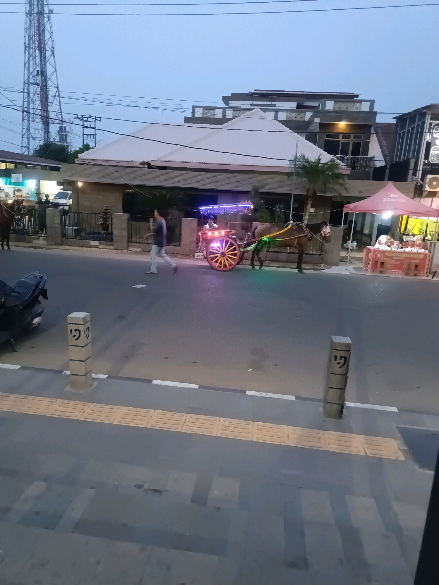 This image depicts a street scene in Lubuklinggau, Indonesia, possibly in the evening or dusk. The foreground shows a paved road with a yellow tactile paving strip along the sidewalk. Two concrete posts with engraved symbols stand on the sidewalk.
In the mid-ground, a horse-drawn carriage with decorative colorful lights is parked on the street. A person is walking away from the carriage towards the left side of the image. A dark-colored motorcycle is parked on the far left. Behind the carriage, there is a stall with a red tent canopy, under which a person appears to be serving customers. Further back, there are buildings with modern architecture, including one with a white roof and balconies, and another with a multi-story structure. A tall transmission tower is visible in the upper left corner of the image. The sky is overcast and a pale blue.