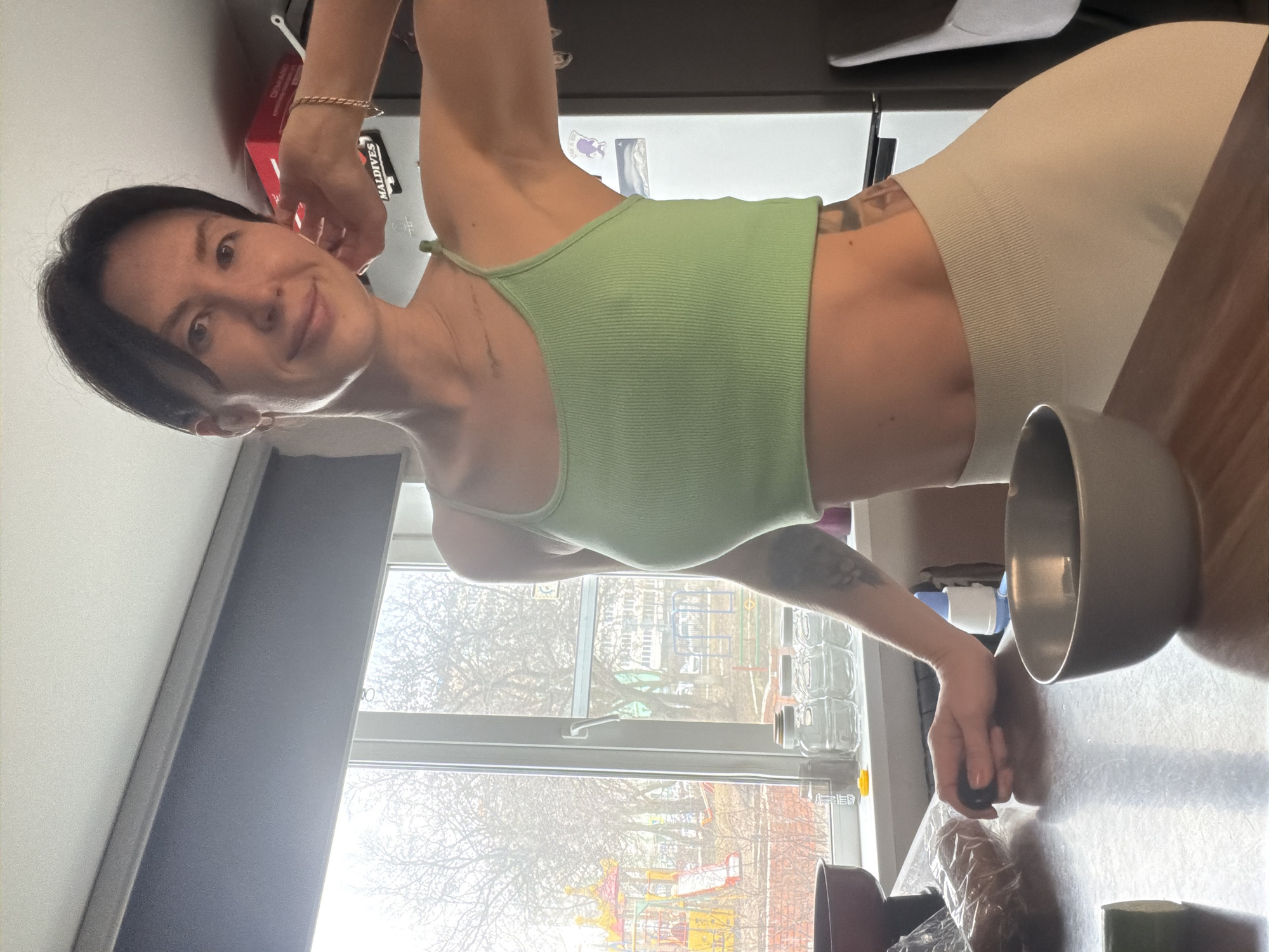 A woman with short dark hair is standing in a kitchen, wearing a green tank top and light-colored leggings. She is smiling and looking at the camera. A gray bowl is on the kitchen counter next to her. A window is visible behind her, showing a playground and a building. The playground appears to be located near Perm, Russia.