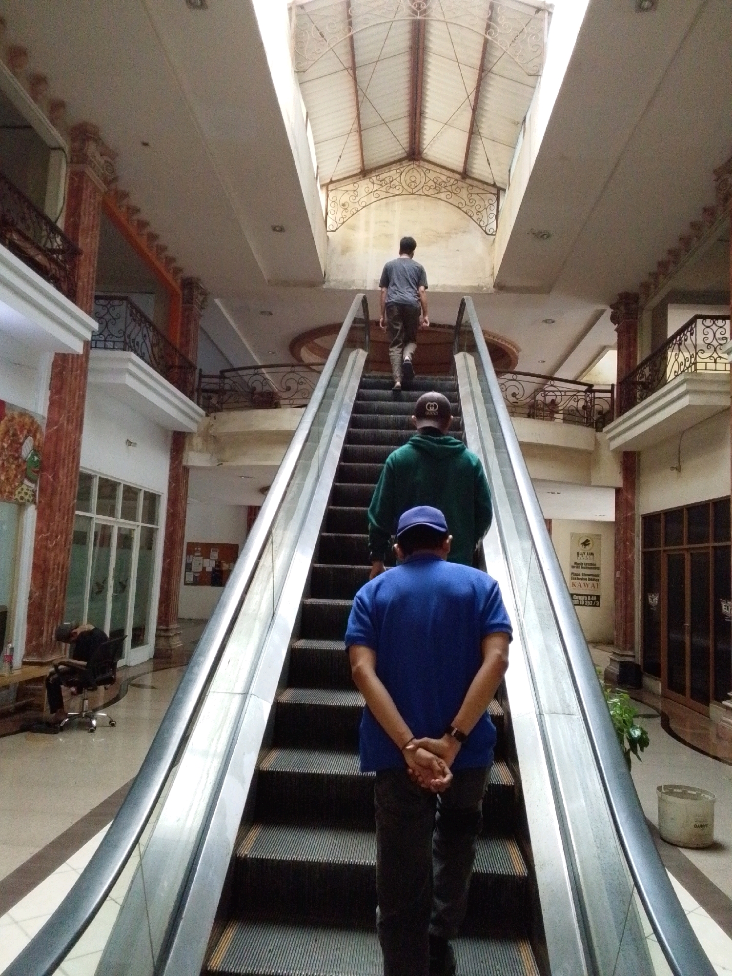 The image depicts an interior view of a multi-story building, possibly a shopping mall or office complex, featuring an escalator. Four individuals are visible, all ascending the escalator.
In the foreground, a person wearing a blue polo shirt and dark pants stands facing away from the camera, with their hands clasped behind their back. To their right, on the escalator steps, is another individual in a green hooded sweatshirt and a baseball cap. Further up the escalator, a third person in a grey t-shirt and dark pants is seen walking. At the very top of the escalator, another person is partially visible, also ascending.
The environment includes polished floors, marble-clad pillars, and ornamental railings on the upper levels. A large skylight is visible at the top of the structure, allowing natural light to illuminate the scene. There are also some architectural details like decorative ironwork and recessed lighting. On the left side of the image, near the bottom, a person is seated in an office chair. Posters and signage are visible on the walls in the background. One sign prominently displays the name "ELLY LIM" and mentions "World's Leading for All Instruments." This detail, coupled with the specific reference to "North Jakarta, Indonesia" in the prompt, suggests the location is likely a commercial or entertainment establishment within that area.