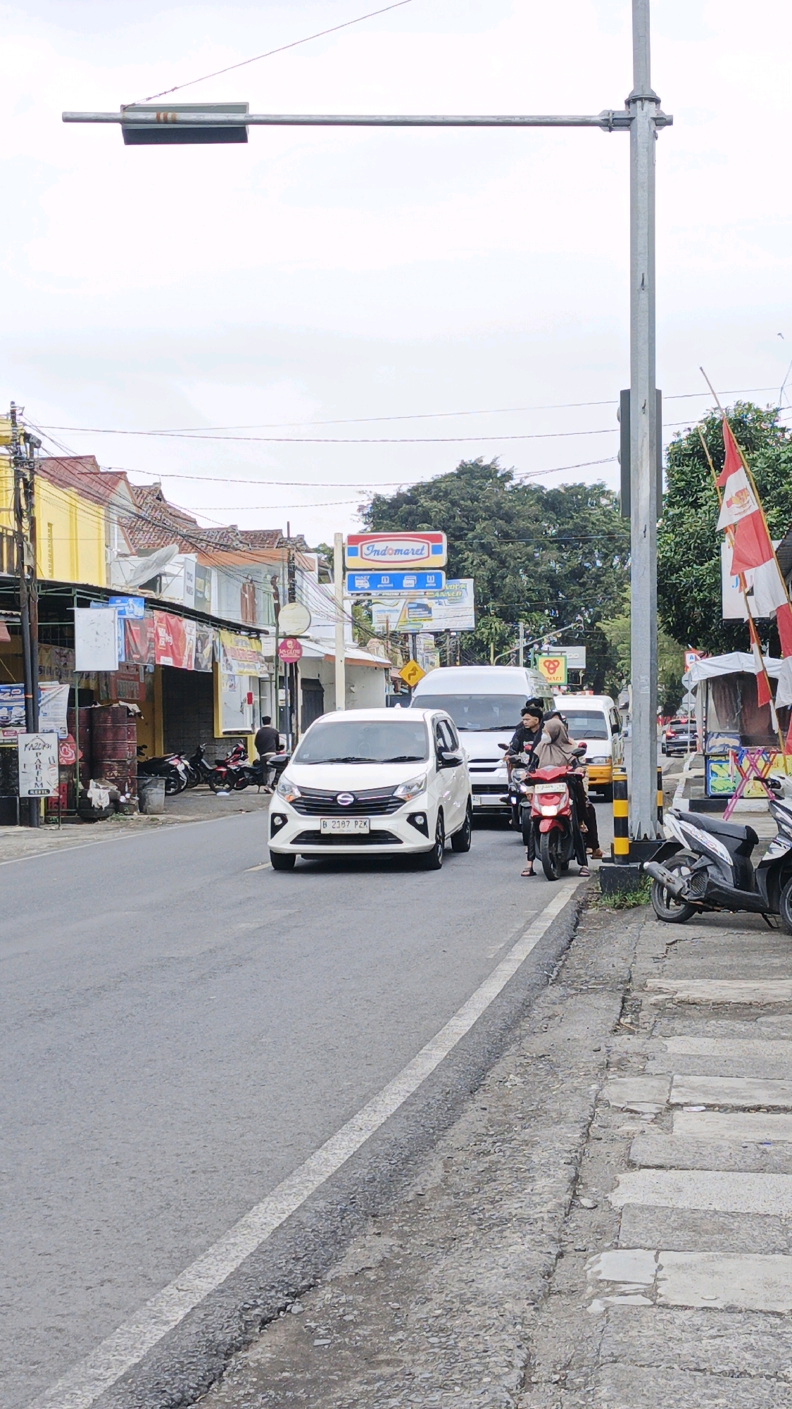 The image captures a street scene in Desa Karangbenda, Indonesia, under an overcast sky. A paved road runs horizontally through the frame, bordered by a gravel and concrete sidewalk on the right and a sidewalk and buildings on the left. Traffic includes a white car in the foreground with license plate B 2187 PZK, a red motorcycle with two occupants, and two white vans further down the road. Several parked motorcycles are visible along the left side of the road, near shops and buildings, some adorned with banners and signs, including an "Indomaret" sign. Flags, one red and white, are displayed on a pole on the right side of the street. The environment appears to be a commercial or mixed-use area with a somewhat weathered infrastructure.