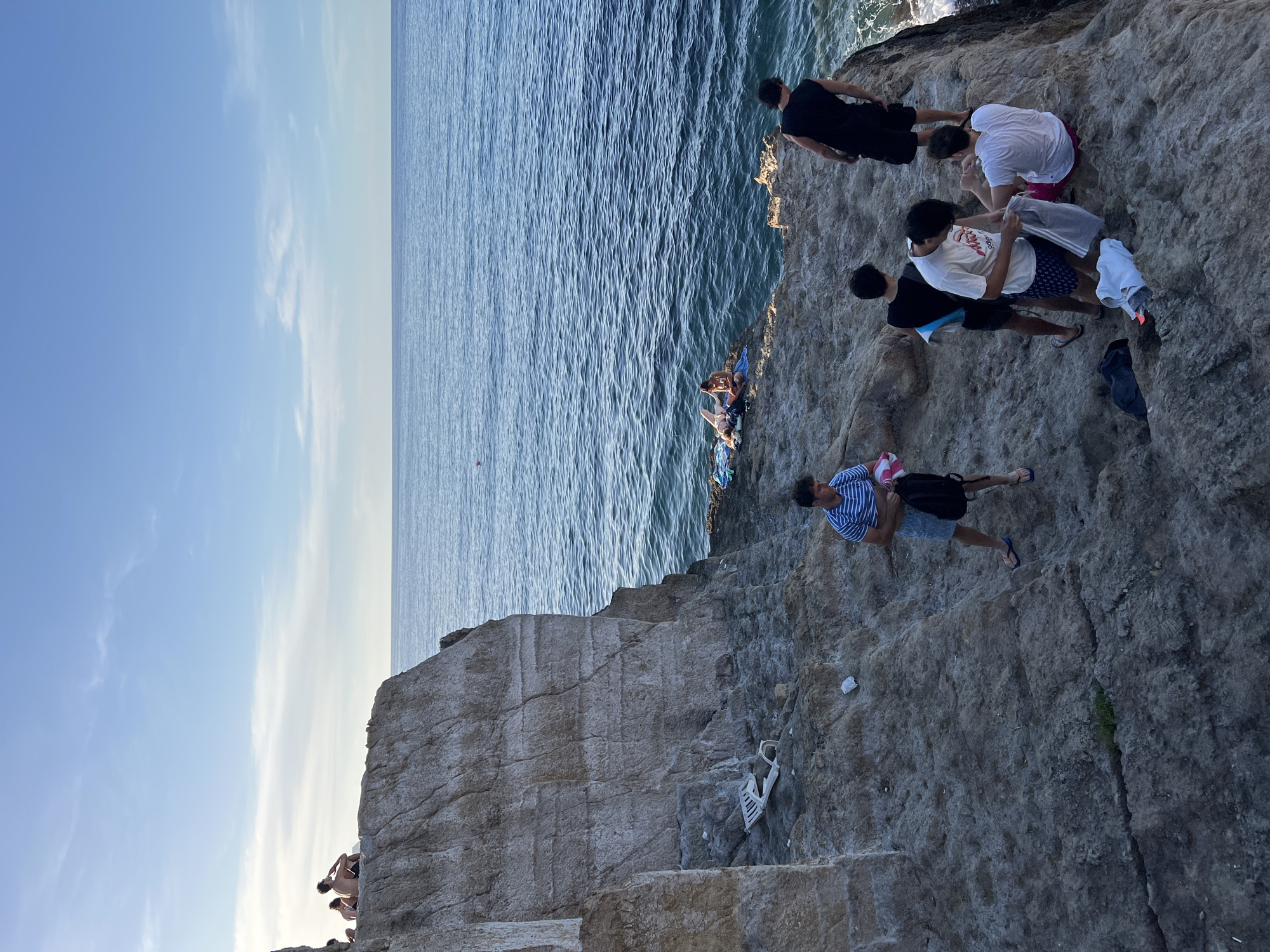 This image depicts a coastal scene in Sperone, Italy, under clear blue skies, likely in the late afternoon. The foreground features a large, light-colored stratified rock cliff on the left, with rough, uneven rocky ground extending towards the right and the sea.
On the upper left edge of the main cliff, two individuals are seated, observing the distant horizon. Further down the cliff face, resting on a small ledge, is an overturned and broken white plastic garden chair, accompanied by scattered small pieces of white litter.
The deep blue sea occupies the right side of the frame, showing visible ripples on its surface. On a lower rocky outcrop by the water, two individuals are relaxing on blue and white towels. One person is reclining, partially obscured by a towel, while another sits upright.
In the lower right section of the image, a group of approximately seven young men is gathered on the rocky terrain. One man in a blue and white striped t-shirt and patterned shorts, wearing blue flip-flops, stands holding a black bag and a pink item. Closer to the water's edge, a man in a black tank top and shorts stands looking out. Another man in a black tank top stands facing inwards. A third man in a white t-shirt with a graphic and patterned shorts is seen holding a grey garment from a clear plastic bag. Seated nearby is a man in a white t-shirt and pink/red shorts, looking towards the individual with the bag. Scattered on the rocks around this group are a black bag, a white towel, and other personal items in plastic bags. A small patch of green vegetation grows in a crack in the rock at the bottom center.