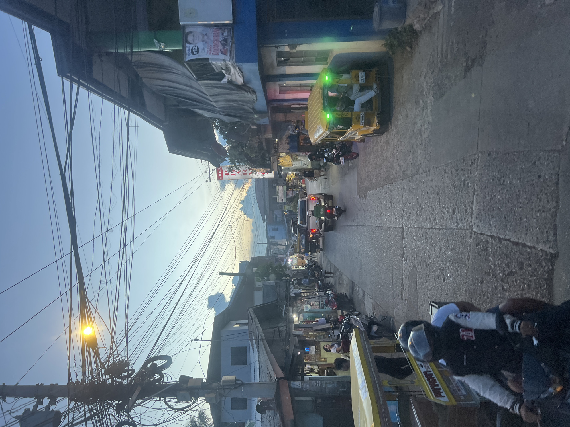The image depicts a street scene in Davao City, Philippines, during either dawn or dusk, as indicated by the soft, diffused light and the twilight sky. The street is paved with cobblestones and is lined with buildings of varying architectural styles, some appearing to be shops or small businesses with awnings and posters. A prominent feature of the urban landscape is the dense network of overhead electrical wires, characteristic of many Philippine cities.
Several vehicles are present on the street, including motorcycles, a yellow jeepney, and a white pickup truck. People are seen riding motorcycles, with at least one passenger wearing a helmet and a jacket. A person is seated on the back of a motorcycle in the foreground. The yellow jeepney, a common mode of public transportation in the Philippines, has passengers inside and is illuminated with green lights. The white pickup truck has the Grab logo on its rear.
A poster with a portrait of a man and the text "Vote! #12 DANNY DAYANGHIRAN" is visible on the facade of a blue building, suggesting it is an election campaign poster. The overall impression is of a bustling, everyday street scene in a developing urban environment.