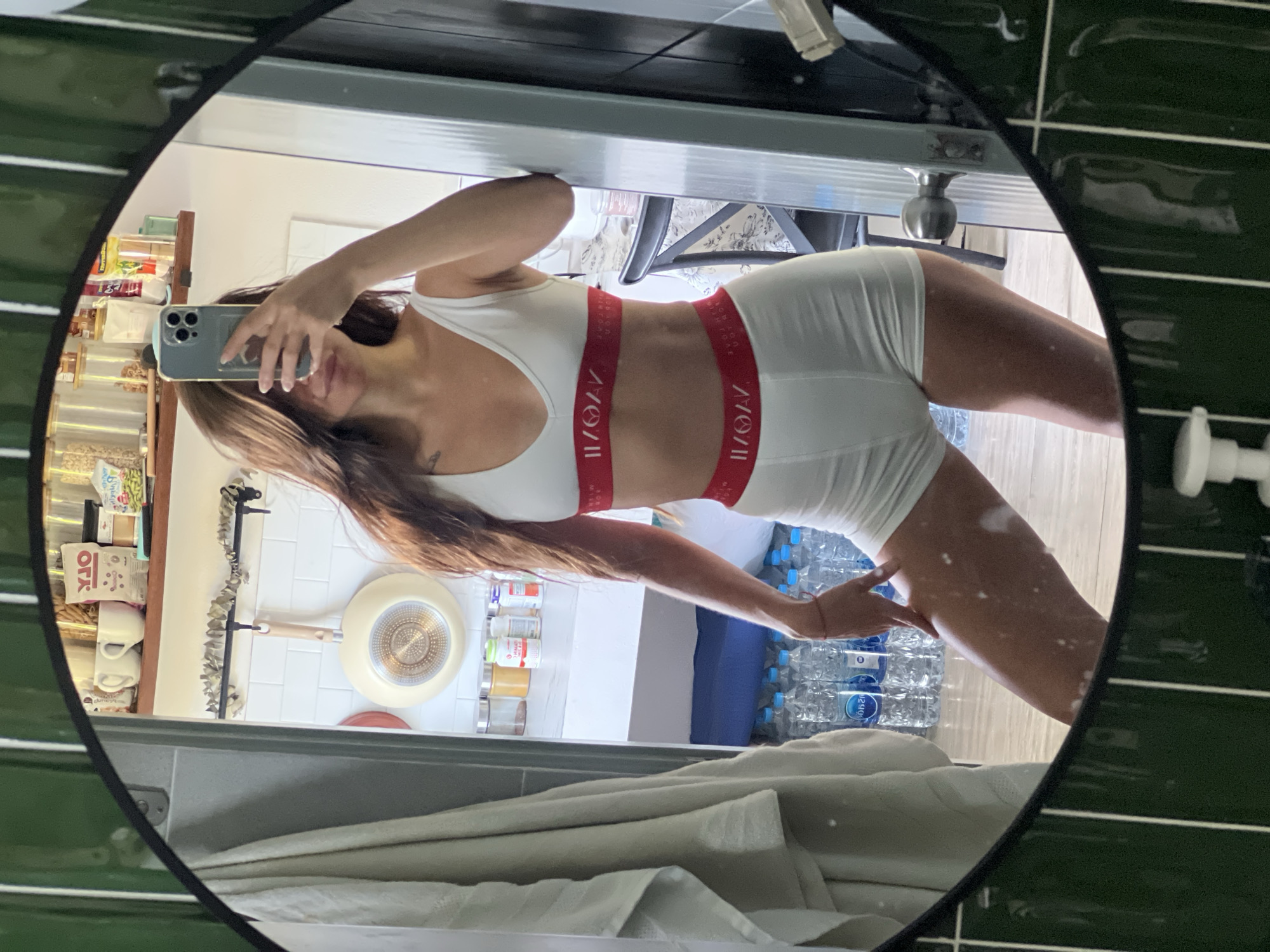 A photograph captured through a circular mirror shows a female figure in a white sports bra and matching shorts with red branding on the waist and chest bands. She is holding a smartphone, presumably to take the picture. The surrounding environment suggests a kitchen space, with visible elements such as storage jars, a cooking pan hanging on the wall, and a shelf with food items. A stack of bottled water is present near the bottom of the frame. The mirror's frame is black, and the background tiles are green.