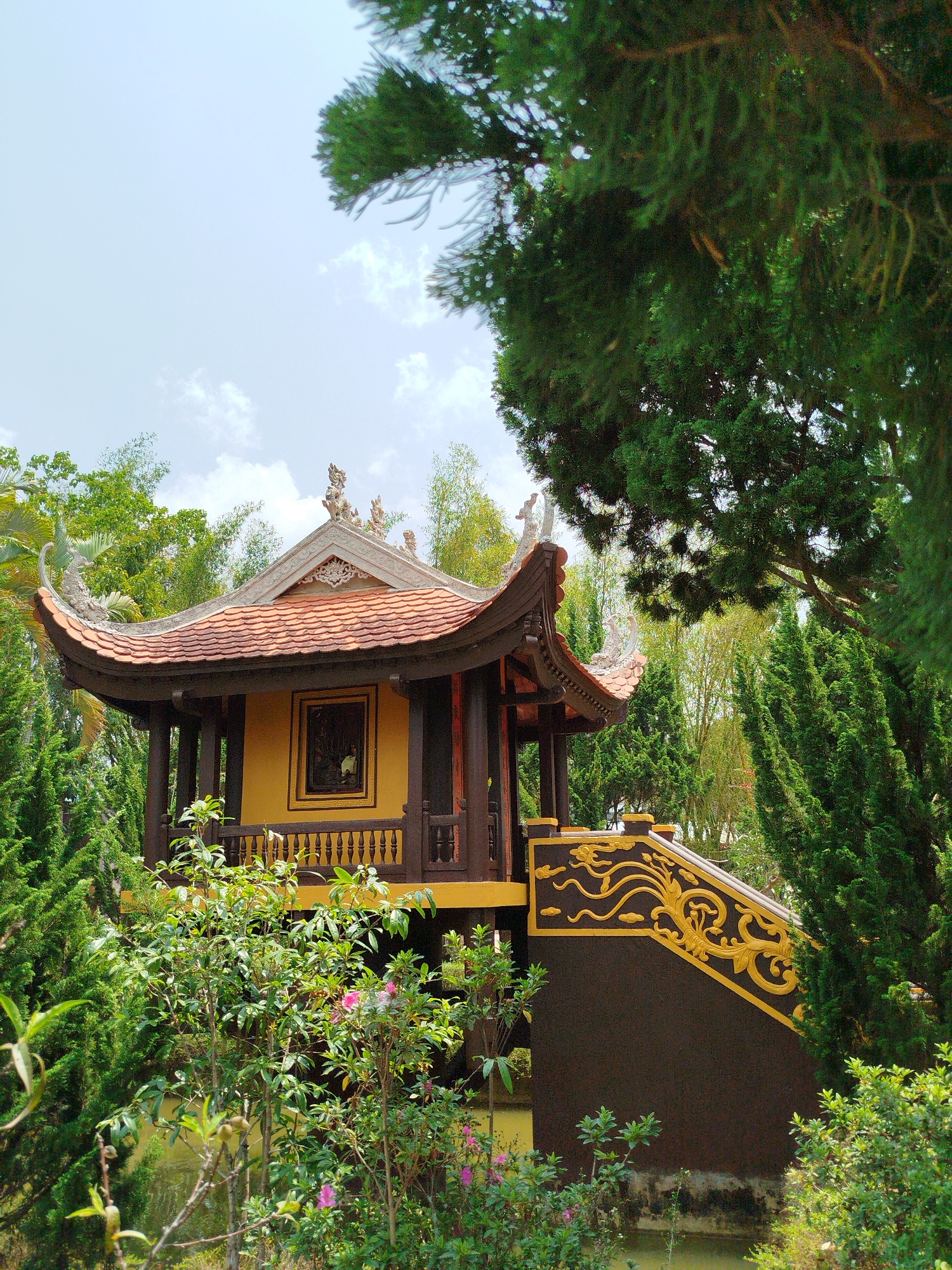 A vibrant photograph showcases a traditional Asian-style pavilion or temple building, nestled amidst lush greenery under a bright, partly cloudy sky. The central structure features a distinctive reddish-orange tiled roof with gracefully curved eaves and ornate white sculptural decorations along its ridges, possibly depicting mythical creatures. The building's walls are a rich yellow, complemented by dark brown wooden pillars, balustrades, and window frames. A framed artwork or carving is visible within a window on the yellow facade.
To the right, a prominent dark brown staircase with intricate gold patterns, featuring swirling and cloud-like motifs, leads up to the pavilion. The entire scene is enveloped by diverse vegetation, including tall, slender cypress-like trees, leafy green trees, and bamboo stalks in the background. In the foreground, various shrubs and bushes, some adorned with small pink blossoms, add to the natural beauty. A body of water or pond is partially visible at the base of the staircase and foreground foliage, reflecting the ambient light.
The image captures a serene, sunlit day, suggesting morning or early afternoon conditions. The architectural style and verdant surroundings are characteristic of a tranquil cultural or religious site in Vietnam, such as a temple or garden within Thôn 5. No people or specific activities are depicted, emphasizing the peaceful and picturesque nature of the location. There is no visible text in the image.
